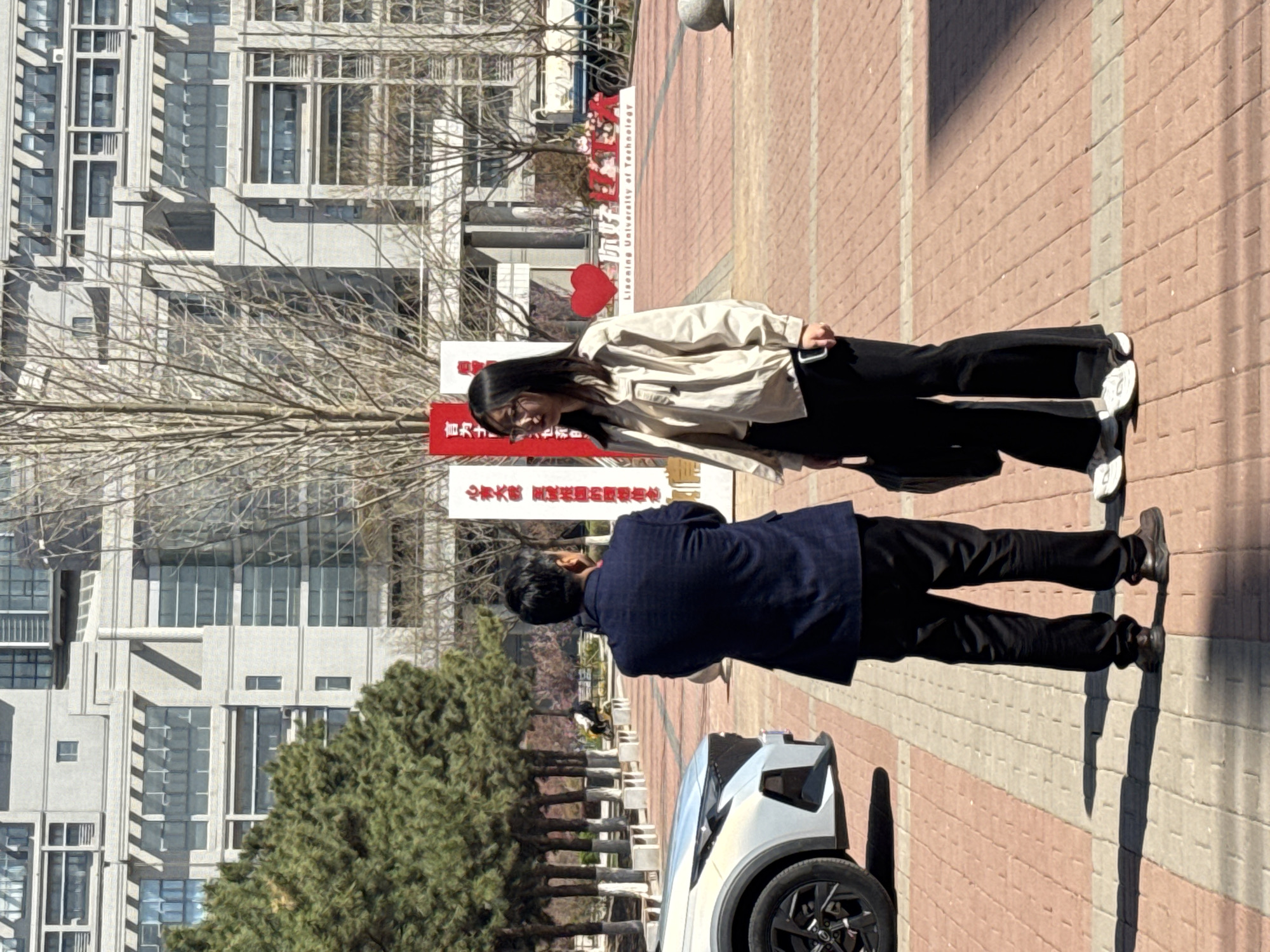 The image shows two people standing on a brick-paved walkway outside a building. The building is likely located at Liaoning University of Technology in Zhongtun, China. A woman is wearing a light jacket and dark pants. A man in a dark blue blazer and dark pants is nearby, standing next to a white car. Behind them are signs with Chinese characters. There are trees and other landscaping in the background. The scene appears to be taking place on a sunny day.