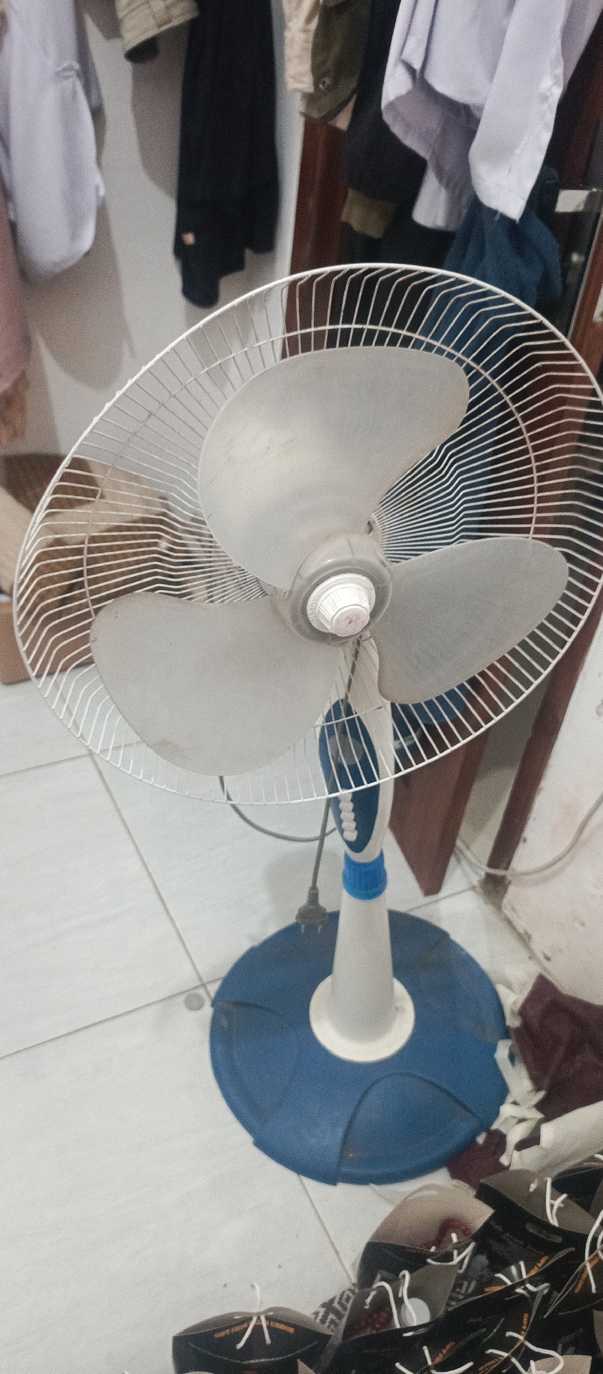 In the city of Arjasa, Indonesia, a stand fan is positioned in an indoor setting, likely a residential area. The fan, predominantly white with blue accents on its base and stand, features a metal grill and three translucent blades. The fan's head is tilted slightly downwards, suggesting it is either in use or positioned for storage. A power cord is visible, plugged into an unseen outlet. In the background, clothing is hanging on what appears to be a wall or a rack, with some garments draped over a wooden frame. The floor is tiled in a light color. The scene appears to be captured during daylight hours, with ambient lighting illuminating the space.