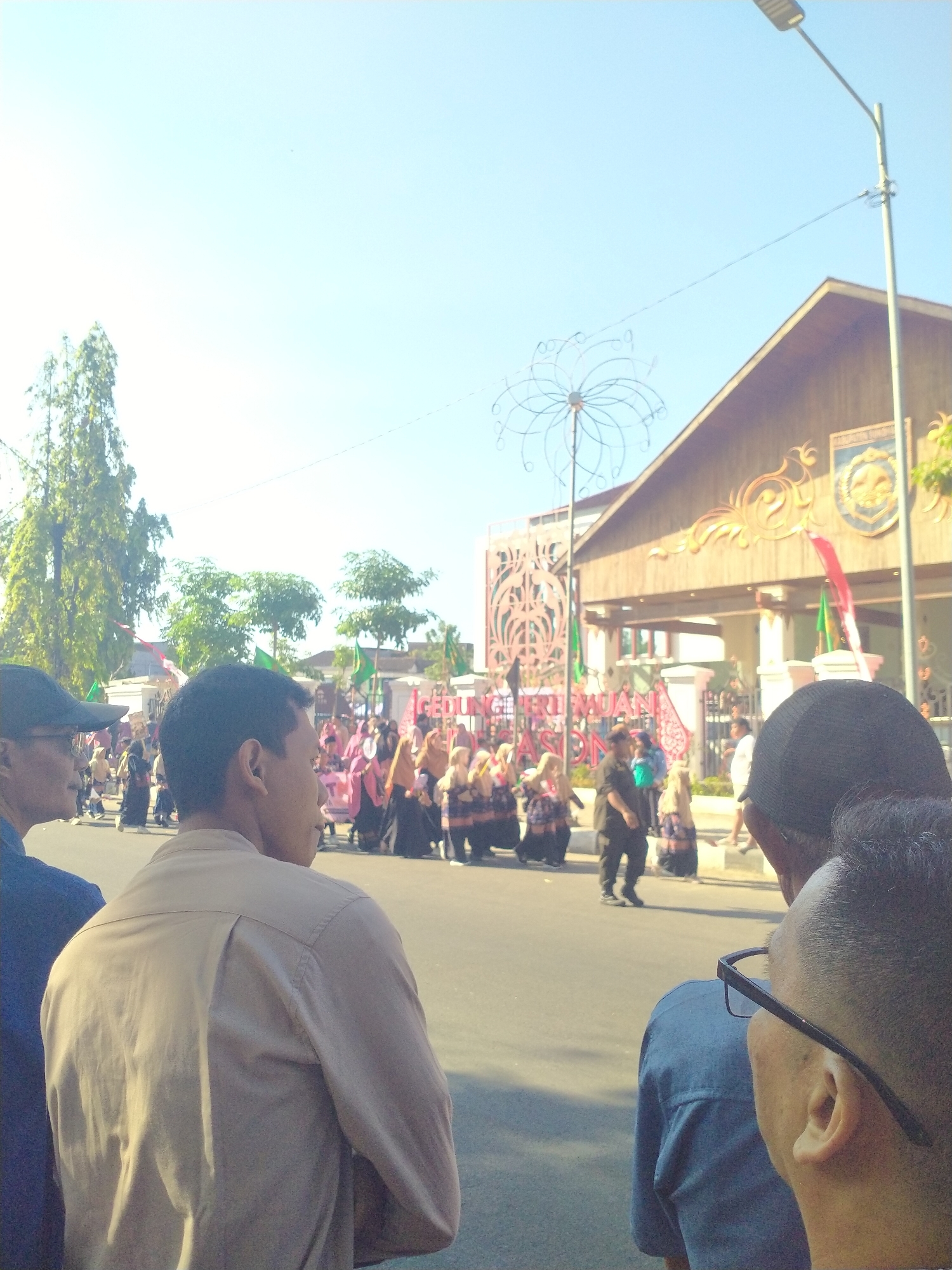 An outdoor, eye-level medium shot shows a street scene in Sukoharjo, Indonesia. In the foreground, two men are visible from the shoulders up, facing away from the camera. The man on the left wears a dark blue shirt and a black baseball cap. The man on the right wears a light brown collared shirt and has short, dark hair. Behind them, a parade or procession is taking place. A group of women, many wearing colorful headscarves and dresses, walk in formation, some carrying flags. To the right, a building with a brown, gabled roof stands prominently. The building has decorative gold trim and a crest or emblem on its facade. A tall street lamp with a single light fixture is visible in the upper right corner. Trees and smaller buildings are visible in the background, and the sky is a clear blue.
