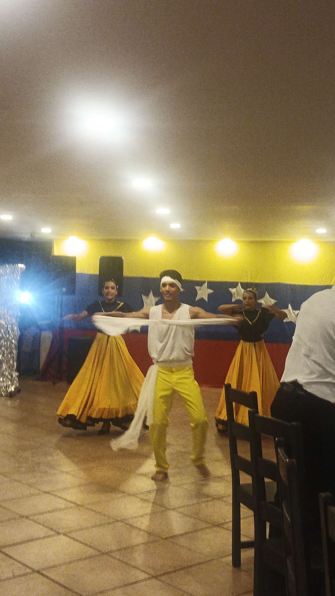 Three people are dancing in a room with a tiled floor. A man in the center wears a white shirt, yellow pants, and a white headband, holding a white cloth extended between his outstretched arms. Two women flank him, each wearing a black top and a long yellow skirt. Behind them is a backdrop painted with the Venezuelan flag's colors and star pattern. To the left, a sequined figure stands partially obscured. A chair is visible to the right of the dancers. The setting appears to be a banquet hall or event space, likely in Playa El Agua, Venezuela, given the backdrop.