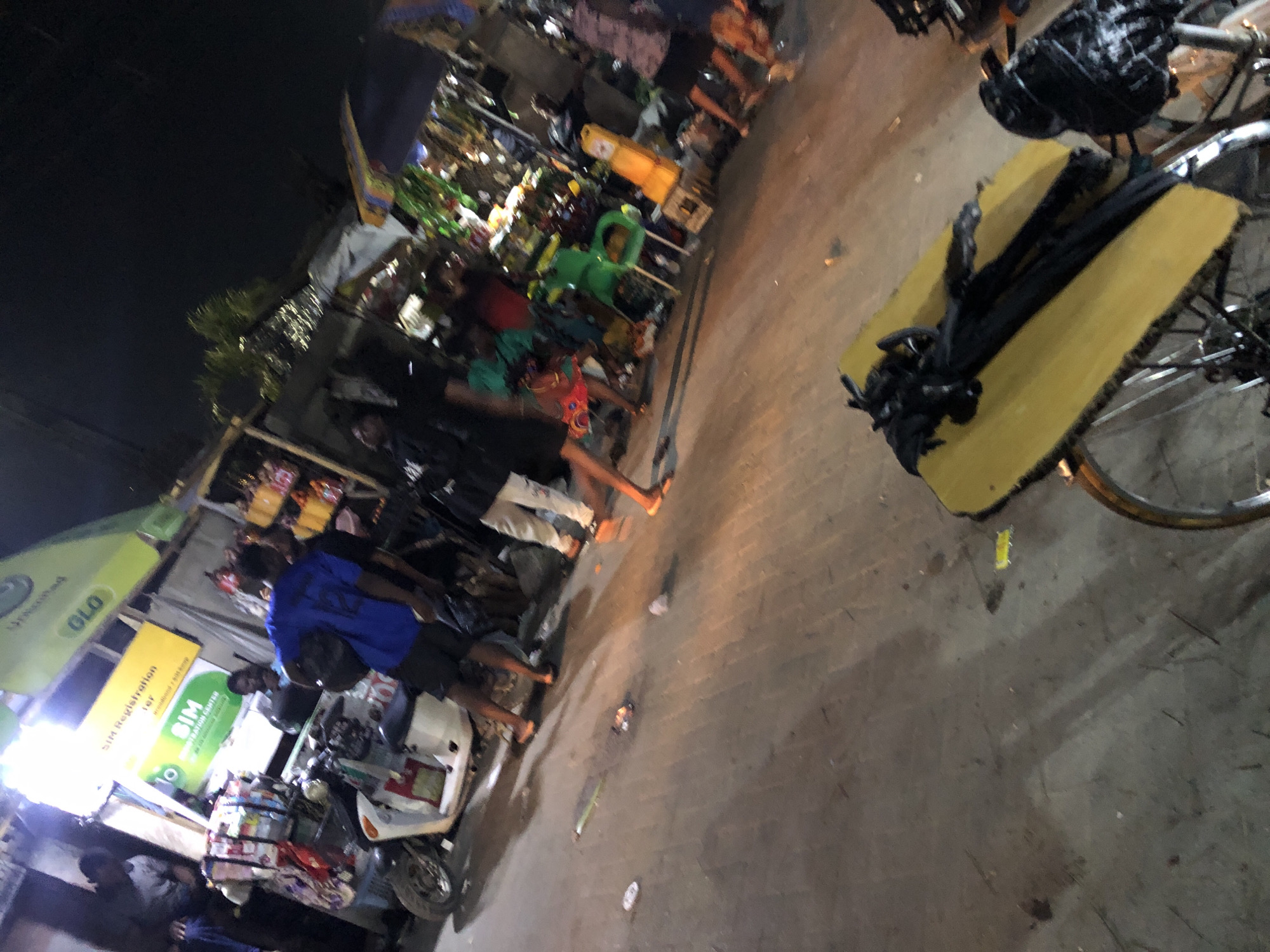 The image depicts a bustling street scene at night in Moba, Nigeria. A small motorcycle is parked near a "SIM Registration" kiosk, next to a "GLO" sign. Several people stand nearby, some barefoot. Merchandise and items clutter the space behind the kiosk. A bicycle with a yellow platform, carrying black fabric or clothing, is positioned prominently in the foreground. The ground appears to be concrete.