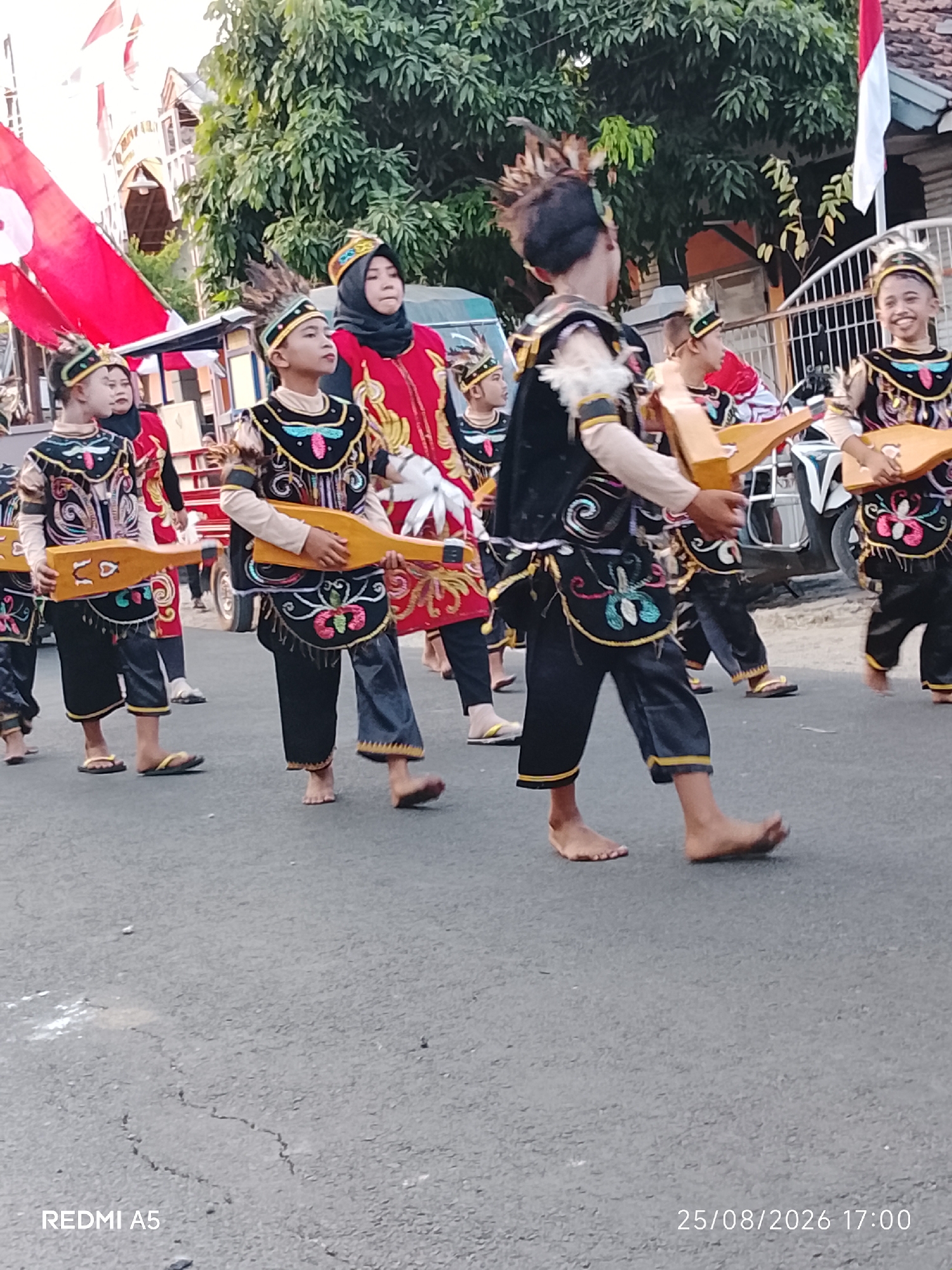 In Semanding, Indonesia, a group of children in traditional attire participate in a procession. They are dressed in black pants and ornately decorated tops with colorful embroidery, headwear with feathers, and some are carrying wooden instruments. A woman in a hijab and red garment walks behind them. They are on a paved road, with trees and buildings visible in the background. A flag is also present. The image is dated August 25, 2026, at 17:00.