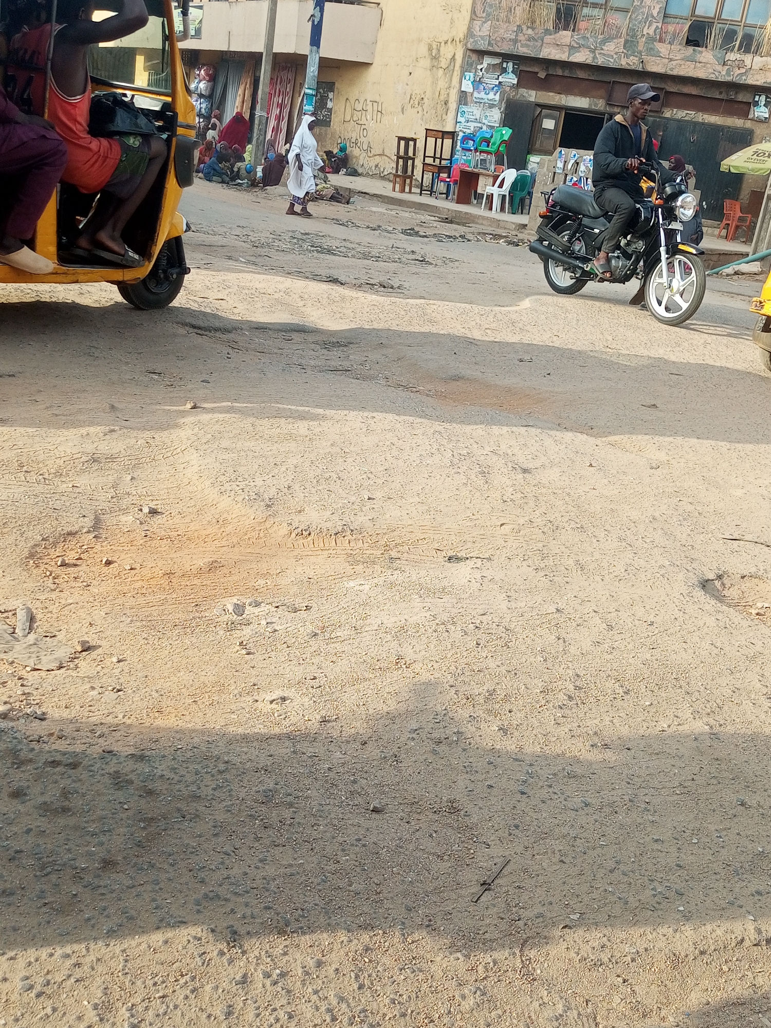 The image shows a street scene in what is reported to be Maiburiji, Nigeria. A yellow auto rickshaw (tuk-tuk) is visible on the left side of the frame, with passengers inside. In the background, several individuals are congregated, some sitting on the ground, and one dressed in white walking. To the right of the frame, a man is riding a black motorcycle. The road surface is dirt and appears uneven, with visible tire tracks. Buildings and other urban elements form the background. A graffiti that says "DEATH TO AMERICA" is visible on a wall in the background.