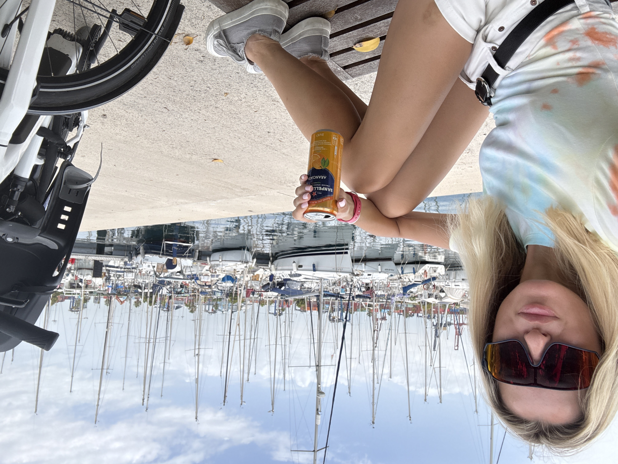 A young blonde woman sits on a wooden bench in front of a marina in Valencia, Spain. She is wearing a tie-dye t-shirt, white shorts, a black belt, and grey sneakers. She is holding a can of San Pellegrino Aranciata. In the background, many sailboats are docked in the marina under a partly cloudy sky. To the right of the woman, the front of a bicycle is visible. The scene appears to be a casual daytime outing.