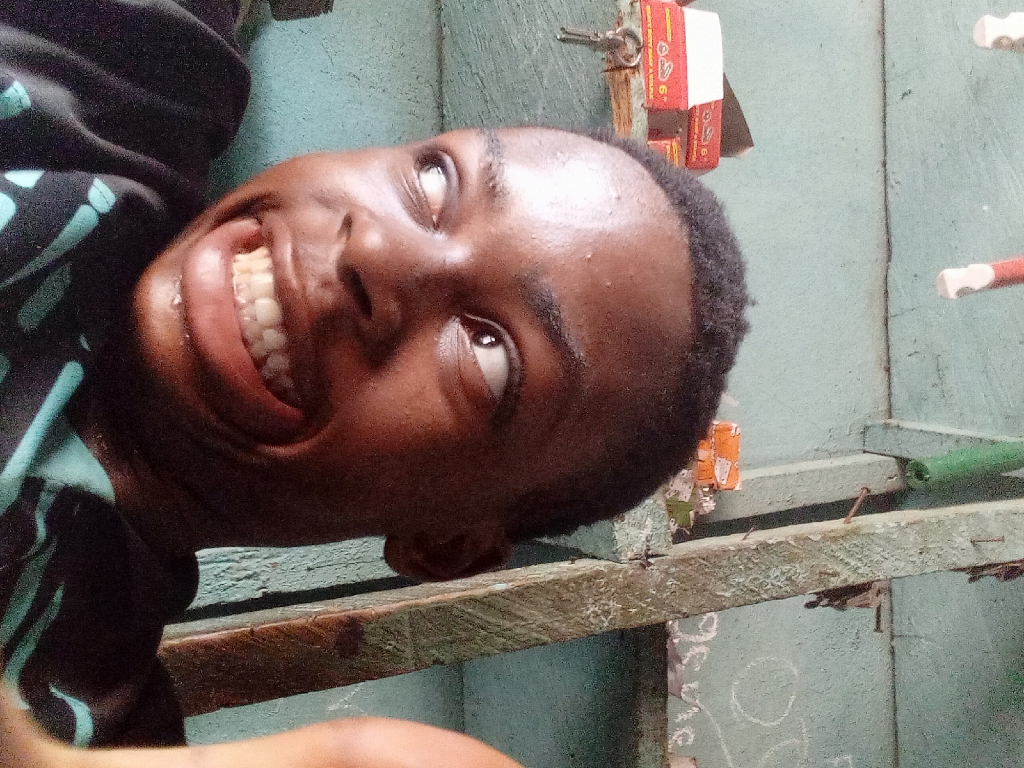 A dark-skinned young man is smiling broadly at the camera. He has short black hair. He is wearing a dark-colored shirt with blue stripes or patterns. The background features a wall with light blue paint, seemingly made of wood. There is a wooden shelf or ledge near the man. There are some objects on the shelf, including boxes. The location may be in Nkpor, Nigeria, as suggested. The overall impression is an informal, close-up shot taken in an indoor environment.