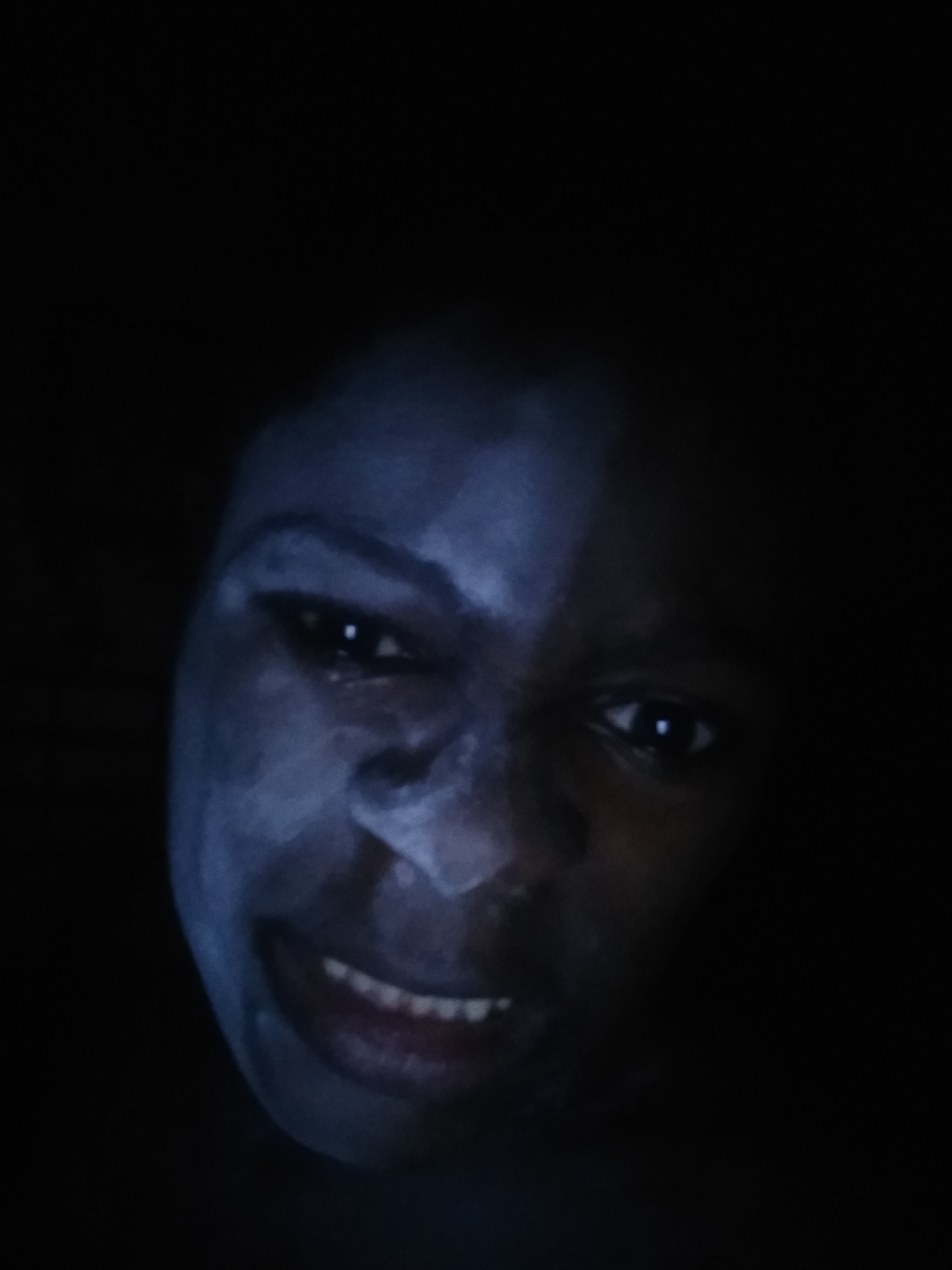 A close-up, low-angle photograph shows a person with dark skin and visible teeth, surrounded by darkness. The person's face appears to have been covered with a pale substance. The surrounding environment is obscured by shadow. The image does not provide any details to confirm the location as Shangisha, Nigeria.