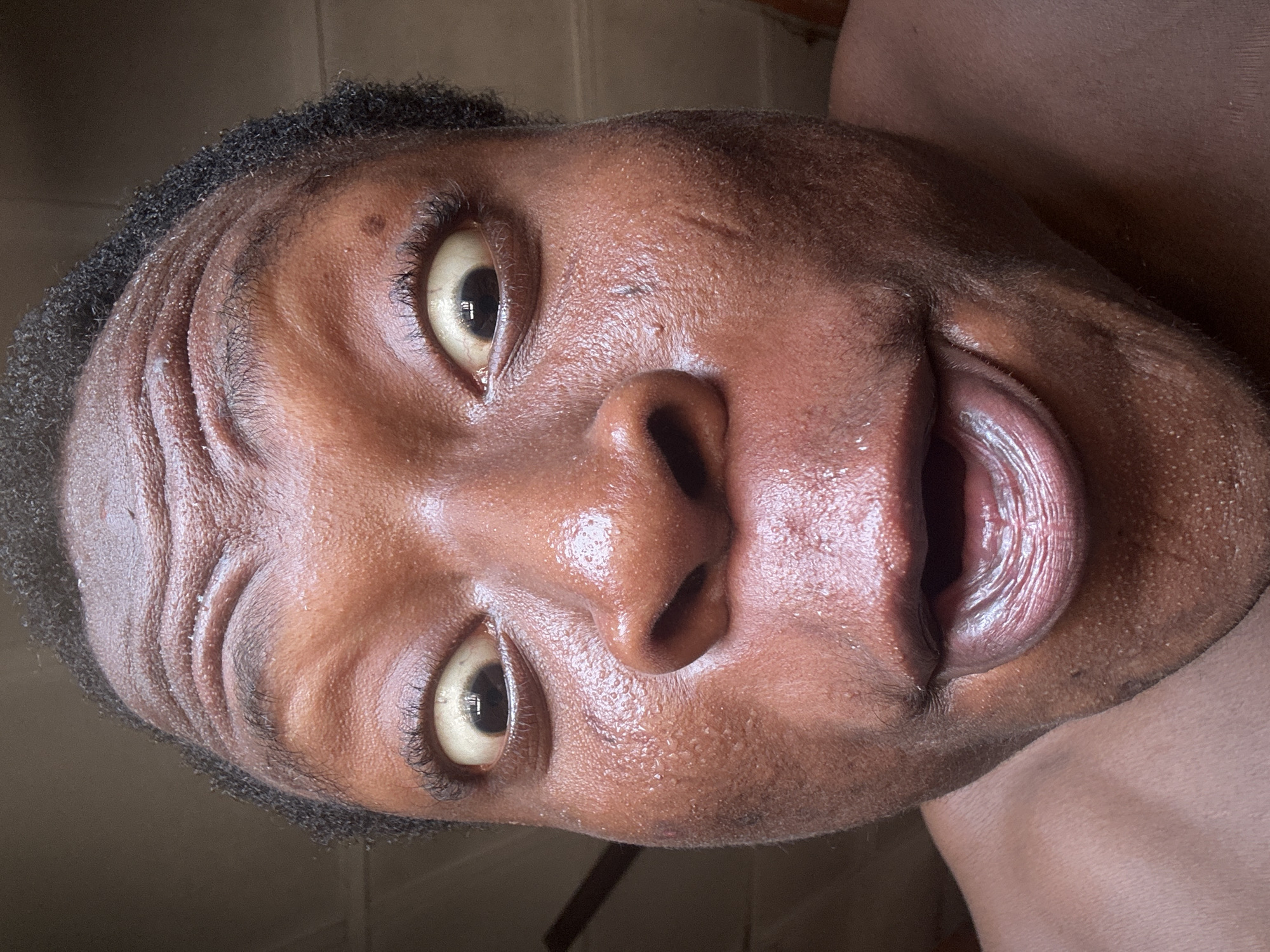 A close-up, upside-down shot of a person's face. The individual has dark skin, black hair, and their mouth is open. Their skin appears to be moist, with visible pores. The background is nondescript. Location is unknown.
