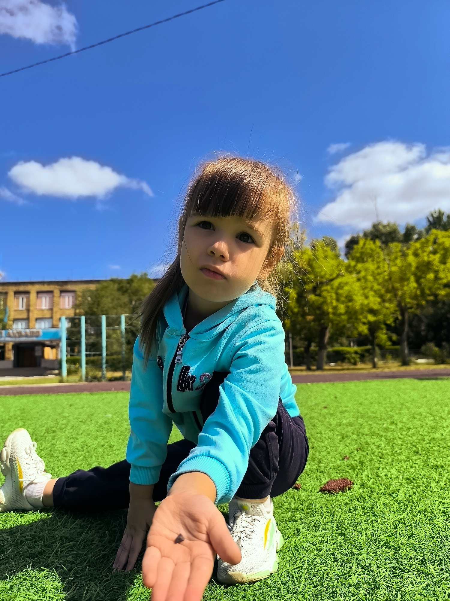 A young girl with auburn hair, wearing a bright blue hooded sweatshirt and dark pants, is crouched on bright green artificial turf. She is holding a small, dark object in her open palm, which is extended towards the camera. Her left leg is stretched out behind her, with her foot in a white sneaker. Her right foot is also visible, positioned in front of her. In the background, a yellow and blue building with multiple windows is visible, along with trees and a blue sky with scattered white clouds. The scene appears to be an outdoor recreational area or sports field within a city.