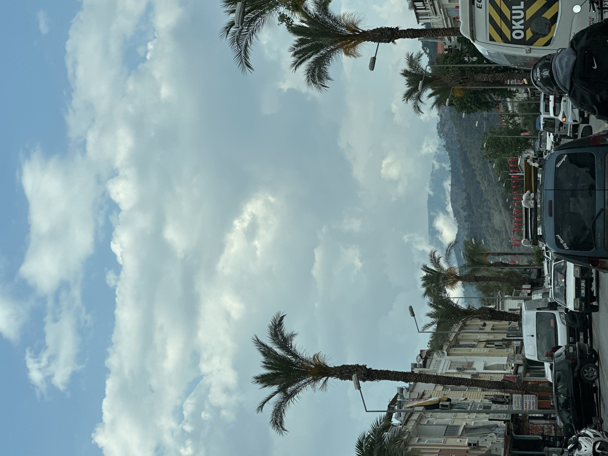 The image shows a street in Fethiye, Turkey, during a cloudy day. Palm trees line the sides of the street, and buildings are visible behind them. A mountain is visible in the distance, partially obscured by clouds. A line of vehicles, including cars, vans, and a school bus, is stuck in traffic. Turkish flags are lined across the street, hanging between the lamp posts. A person wearing a helmet is partially visible in the foreground. Several brands of vehicles are identifiable including FIAT and DACIA.