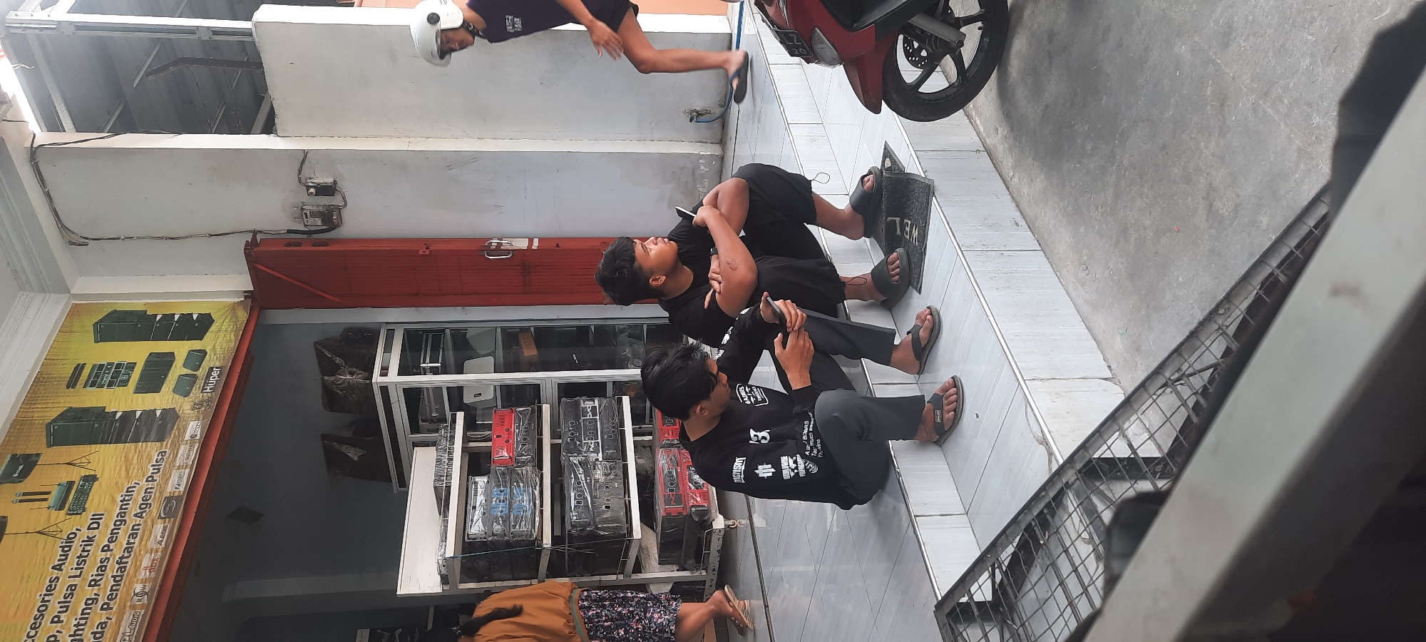 In Selopuro, Indonesia, two young men are seated on the white tiled steps of a storefront. The man on the left, wearing a black long-sleeved shirt and grey pants, is looking at his phone. The man on the right, in a black t-shirt and black shorts, is looking to his left. A third individual, wearing a white helmet, a purple t-shirt, and black shorts, walks away from the viewer on the right side of the frame. In the background, merchandise, including audio equipment and speakers, is visible through the open doorway of a shop. A red motorcycle is parked to the right of the steps. The environment appears to be an outdoor commercial area with a concrete ground.