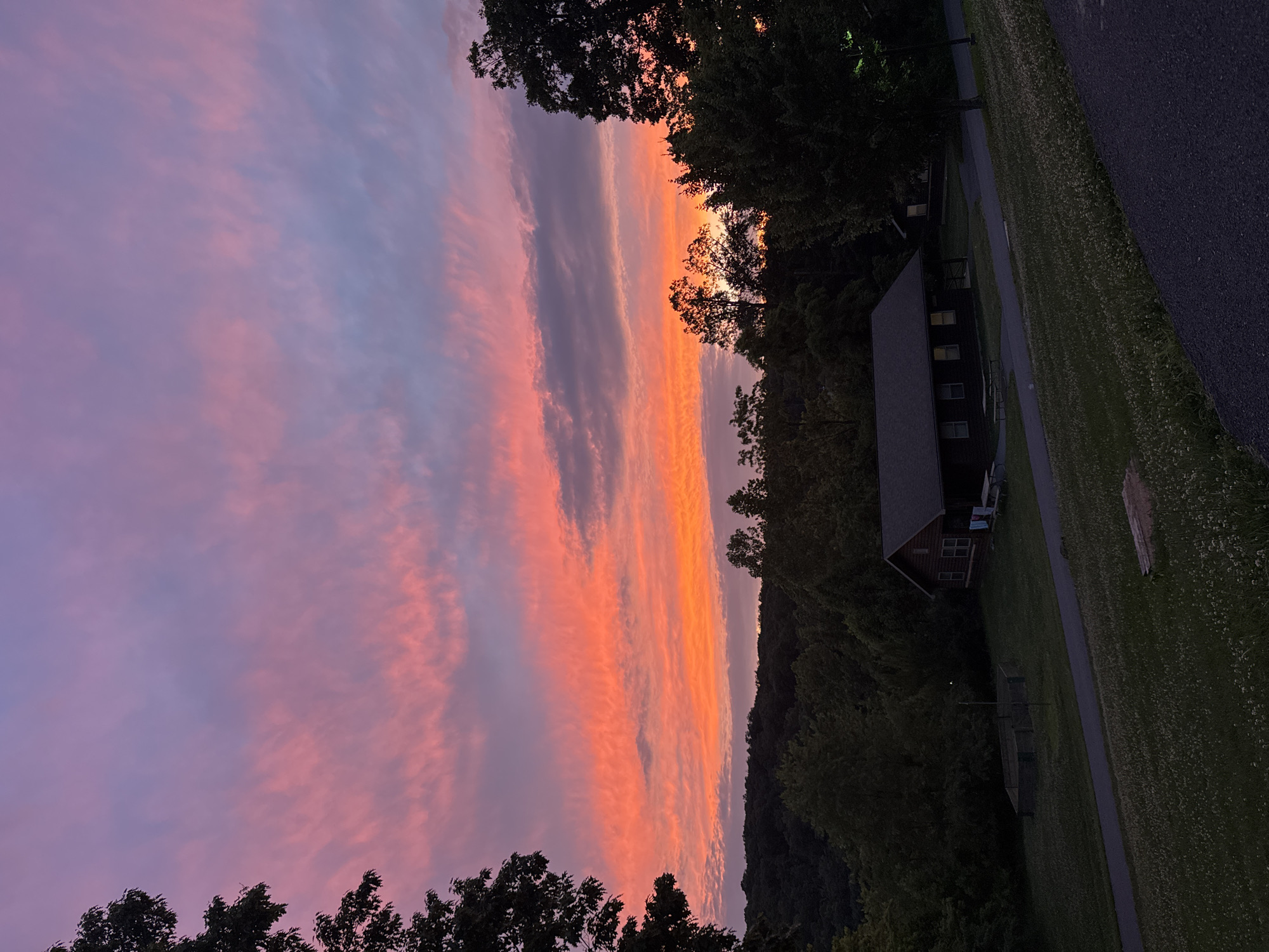 This is a horizontal, eye-level, medium shot of a scenic sunset over a wooded area with a few buildings. The sky is filled with vibrant streaks of pink, orange, and purple clouds. A silhouette of trees lines the horizon, with a dark forested area behind them. In the foreground, there are two small, rustic cabins situated on a grassy lawn. One cabin, larger than the other, has a dark roof and a few windows visible. A picnic table sits in front of it. A paved path runs through the scene, and a small, fenced-in area is visible to the left of the larger cabin. The overall impression is peaceful and natural.