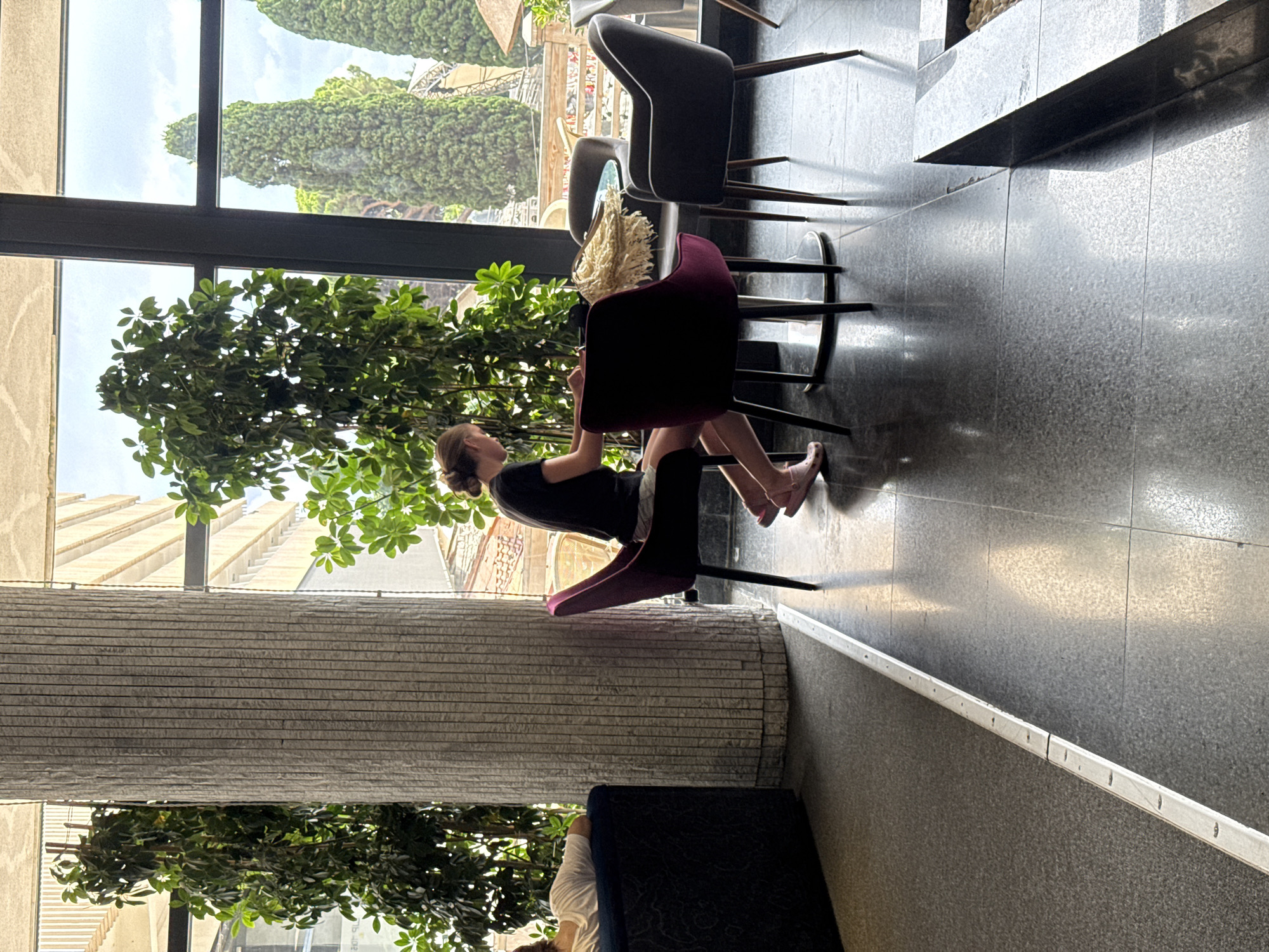 A young woman with blonde hair pulled back is seated at a round table in a brightly lit indoor space. She is wearing a black t-shirt and light-colored shorts, with pink slip-on shoes. Her focus is directed downwards, towards items on the table in front of her. To her right, a woven item, possibly a placemat or basket, rests on the table. She is seated in a dark red chair. To her left, a dark gray chair is positioned at the same table. Beyond the table, through a large window, lush green trees are visible, with cypress trees and a building in the distance, suggesting an outdoor environment. The interior features a large, textured grey pillar and a dark floor with a visible seam. To the far left, a person is reclined on a dark blue sofa. The overall impression is that of a seating area within a hotel lobby or cafe, with a view of an exterior landscape. The location is identified as a city in Ukraine.