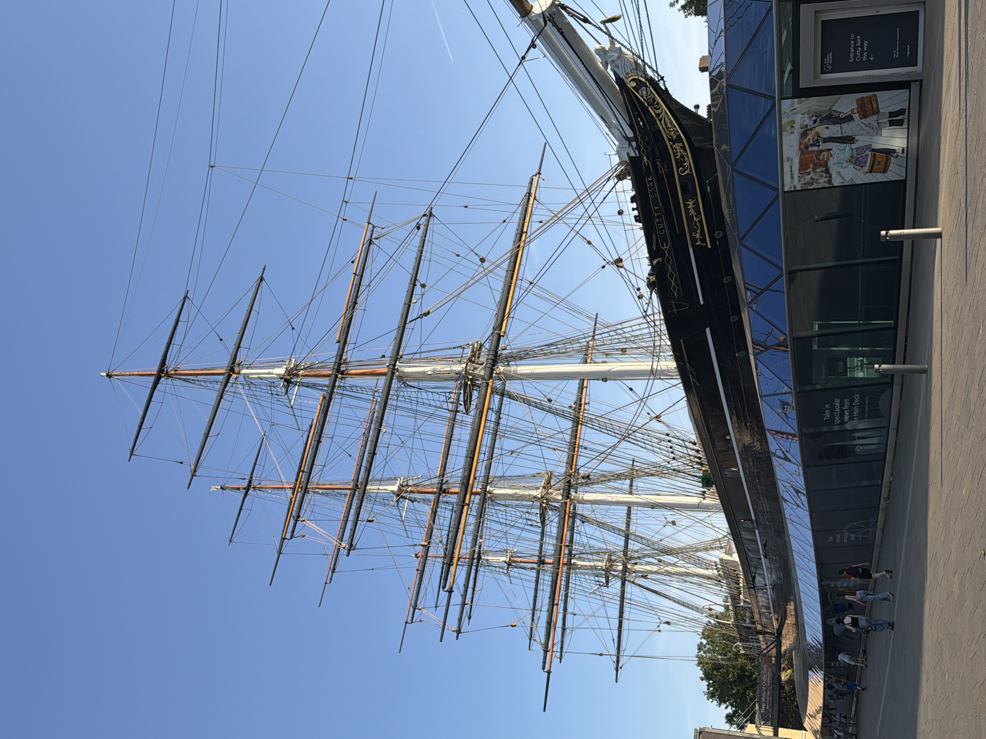 The image shows the Cutty Sark, a British clipper ship, with its masts and rigging prominently displayed against a clear blue sky. The ship's hull, painted black with gold accents, is partially visible alongside a modern building with reflective glass panels. The building has signage related to the Cutty Sark, including directions to the entrance and advertising for the main deck views. Several people are walking along the pavement adjacent to the building and the ship. A few trees are seen near the base of the building. The image is taken from a low angle, emphasizing the height of the ship's masts. The location is most likely Greenwich, London, United Kingdom, where the Cutty Sark is a preserved historical attraction.
