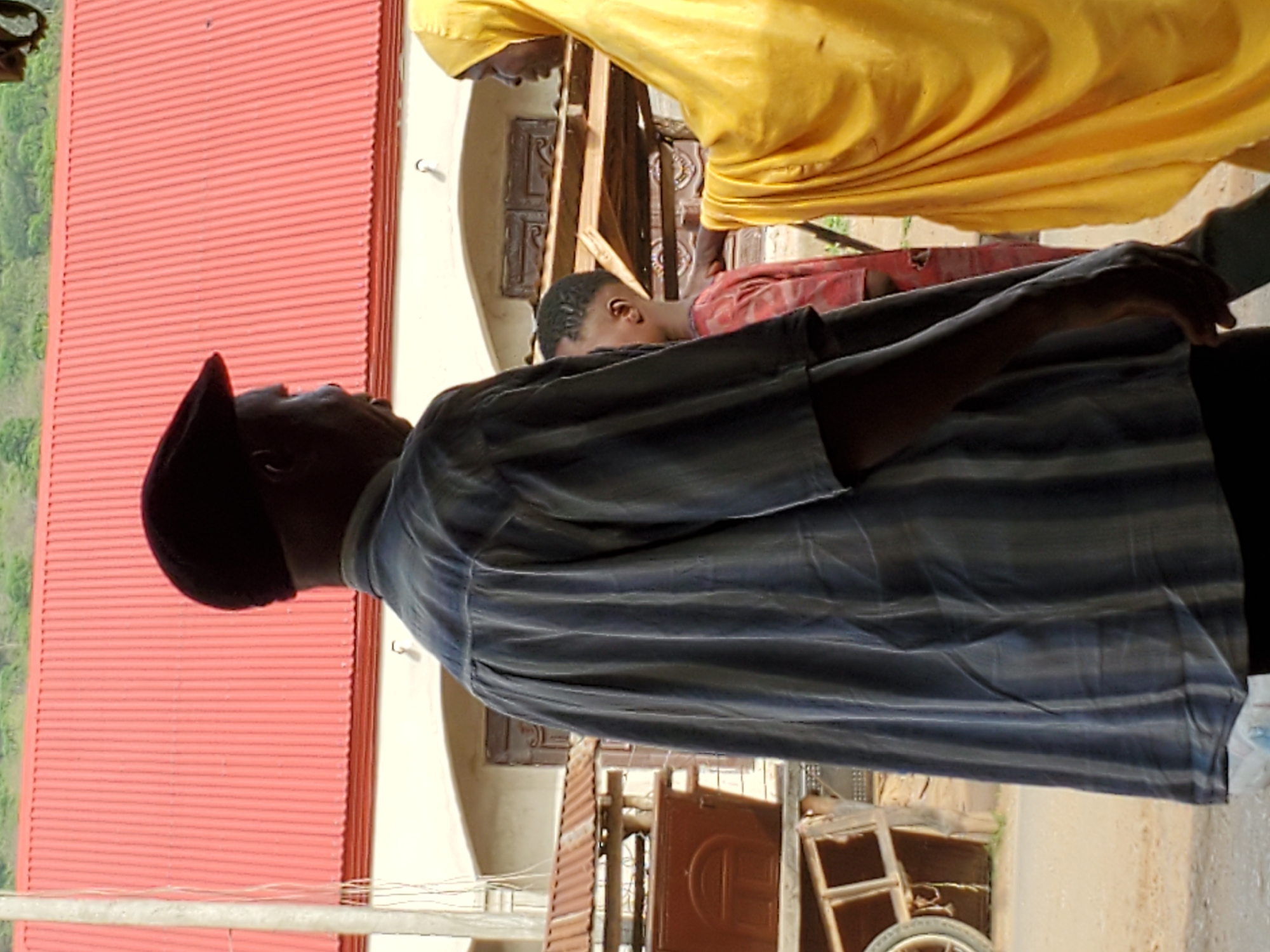 The image, set in Mararaba, Nigeria, captures an outdoor daytime scene featuring three individuals. In the foreground, a dark-skinned man is seen from behind, wearing a dark cap and a light blue and grey striped short-sleeved shirt. He appears to be looking upwards or forward, with his right arm bent at the elbow.
To the right, another dark-skinned person is partially visible, draped in a flowing yellow garment, possibly a traditional robe. This individual's head and upper body are seen, looking downwards. A dark-skinned child, wearing a red top, is partially obscured behind the yellow-clad person, also looking downwards.
The background on the left features a prominent red corrugated metal roof, indicative of a building structure. Behind the people, light-colored walls are visible, along with various wooden architectural elements, possibly doorframes or window frames. Towards the lower right, a wooden structure resembling a cart or stand, equipped with a wheel, is present. The bright lighting suggests a sunny day. No visible text is present in the image.