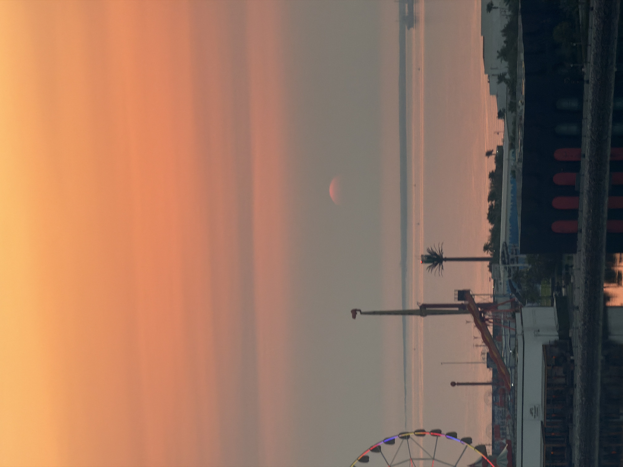 The image depicts a waterfront amusement park at sunrise or sunset. The sky is a gradient of orange and pink hues. A Ferris wheel with illuminated cabins is visible on the left side of the frame. In the center, a tall ride with a red slide mechanism stands prominently. A palm tree is also visible in the distance. The water in the foreground reflects the colors of the sky. Buildings, some with dark facades and arched entrances, line the waterfront, suggesting a commercial or recreational area. A large ship is visible on the horizon, and smaller boats are scattered across the water. The overall scene conveys a sense of a coastal amusement park at dawn or dusk.