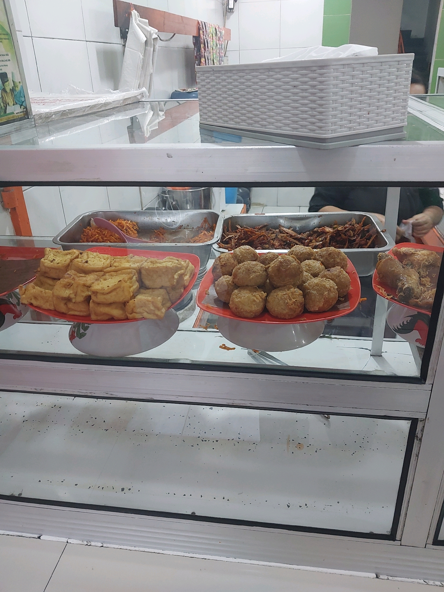 The image shows a display of fried foods in a glass-fronted cabinet, likely in a food stall or small eatery. On the left, a red rectangular plate holds several pieces of golden-brown fried tofu, stacked neatly. To the right of the tofu, another red plate contains a pile of round, breaded fried items, possibly meatballs or croquettes, also golden-brown. Behind these plates, in a metal tray, is a portion of shredded, stir-fried carrots, appearing orange and slightly wilted. Further to the right, another red plate displays a piece of fried chicken with a glaze, alongside some shredded bits that might be vegetables or other fried components. The food is displayed under fluorescent lighting. The background includes a white tiled wall and a wooden shelf with hanging white paper goods. The overall impression is of a self-service or counter-service food vendor, typical of many locations in cities like Malang, Indonesia, where such preparations are common.