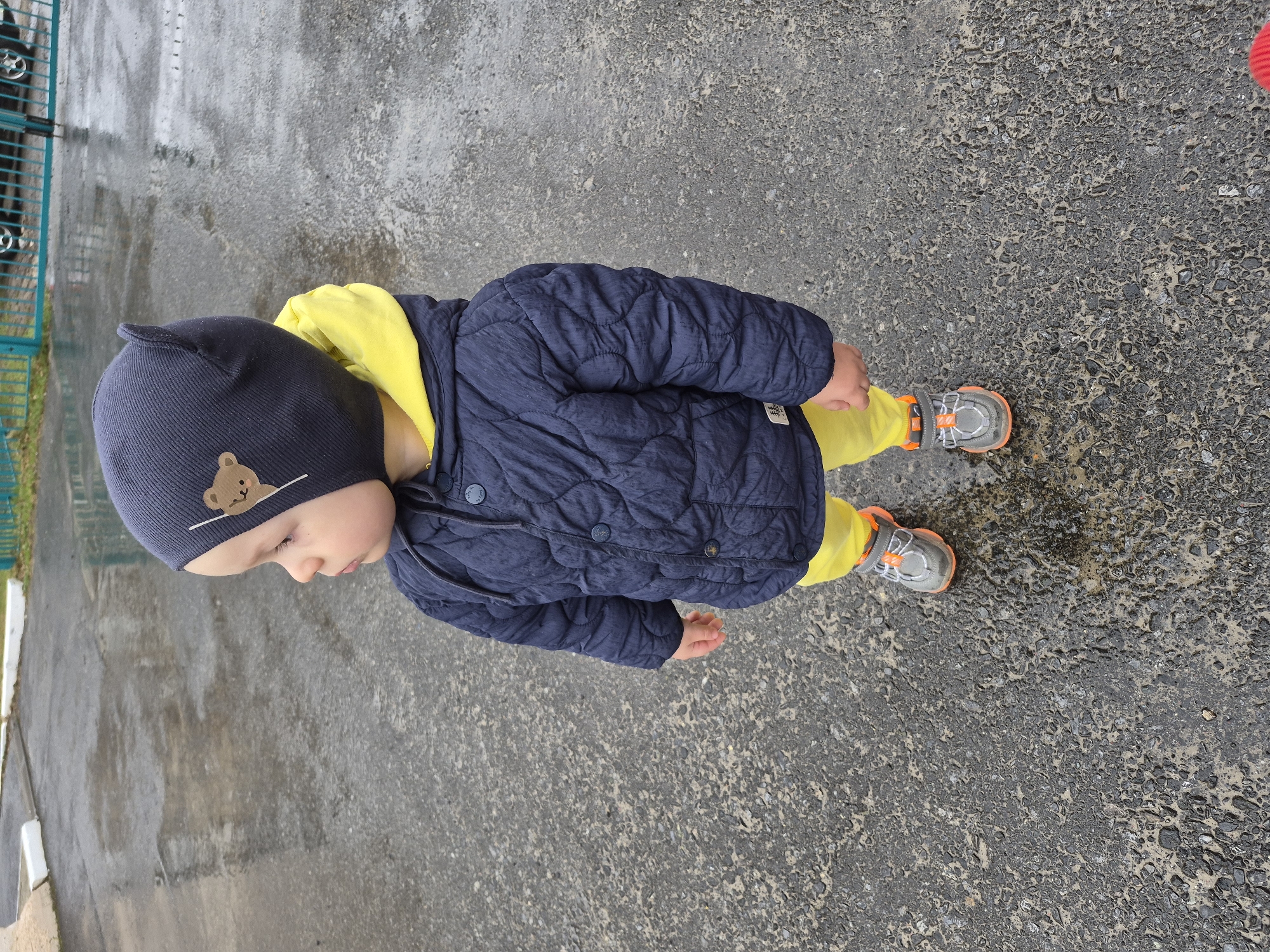 A young child stands on a wet, paved surface, likely a street or sidewalk. The child wears a navy blue quilted jacket, a bright yellow hooded top, yellow pants, and a navy blue hat with a bear design. The child is also wearing gray sneakers with orange accents. The setting appears to be outdoors on a cloudy day, based on the wet ground and lighting. The location is specified as Tobolsk, Russia. A fence with green and white elements is visible in the background on the left side of the image.