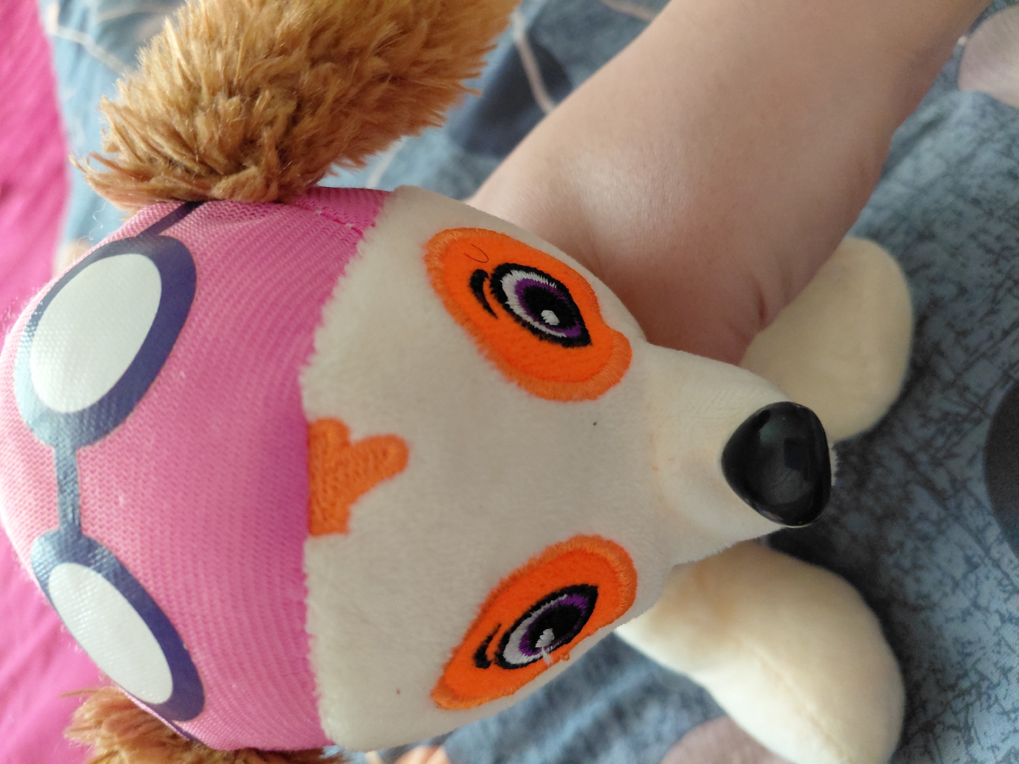 A plush toy, resembling the character Skye from Paw Patrol, is being held by a person. The toy is predominantly white with pink fabric on its head, adorned with purple and white eye graphics, simulating goggles. Orange accents are visible on the toy's face, around the eyes, and as a nose marking. Two brown, furry ear-like appendages protrude from the sides of the head. The toy's body is soft and white, with a black, glossy nose. The background consists of a textured blue fabric and a bright pink surface. The image was captured in Kenzhe, Russia.