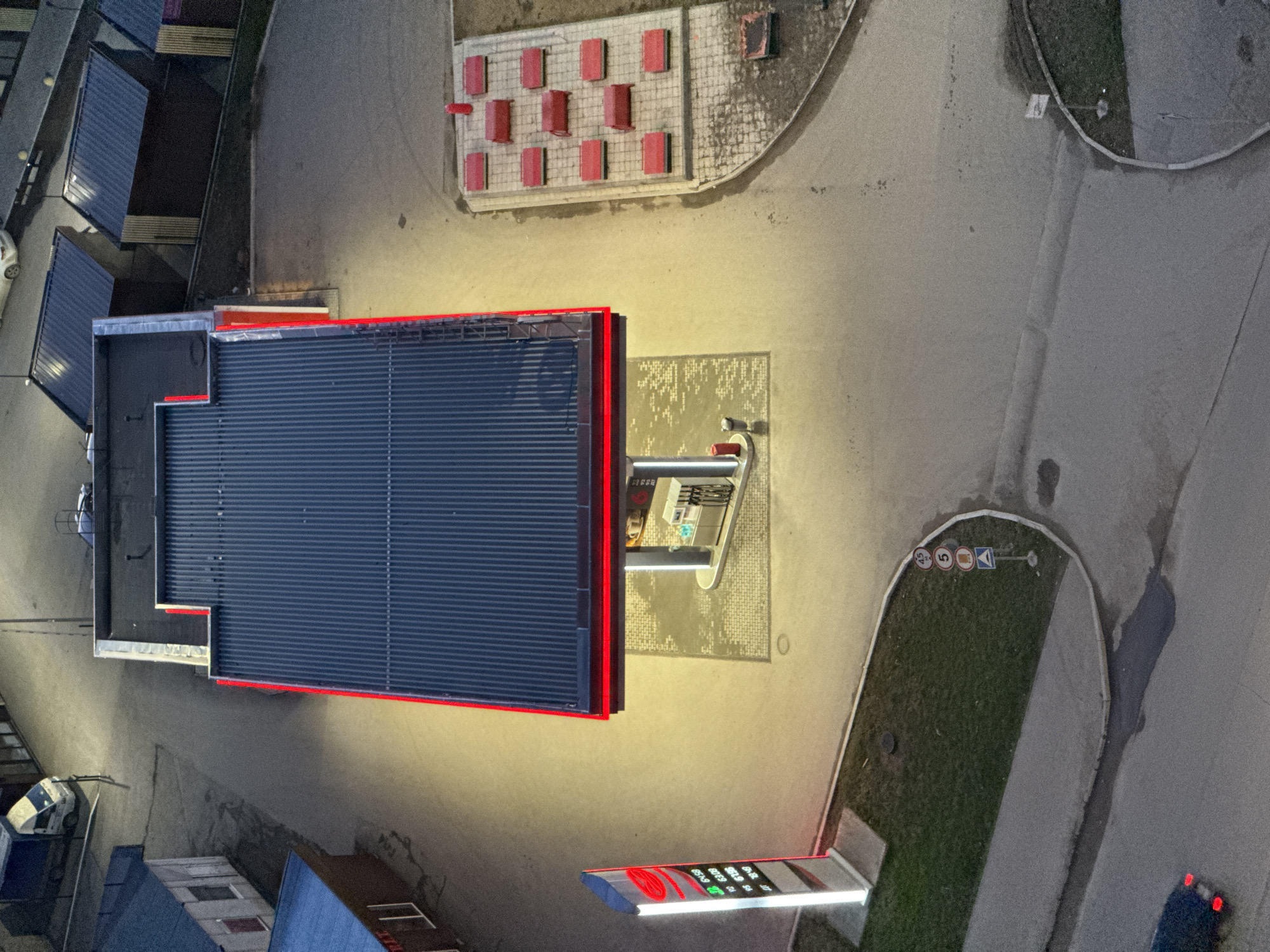 This is an aerial nighttime view of a gas station in Perm, Russia. The gas station has a dark blue corrugated metal roof, a red trim, and illuminated gas pumps. There's a lighted sign displaying gas prices nearby. Adjacent to the gas station is a paved area with red block seating. Nearby are sections of road, grass, and sidewalks, with a few vehicles and buildings in the surrounding area.