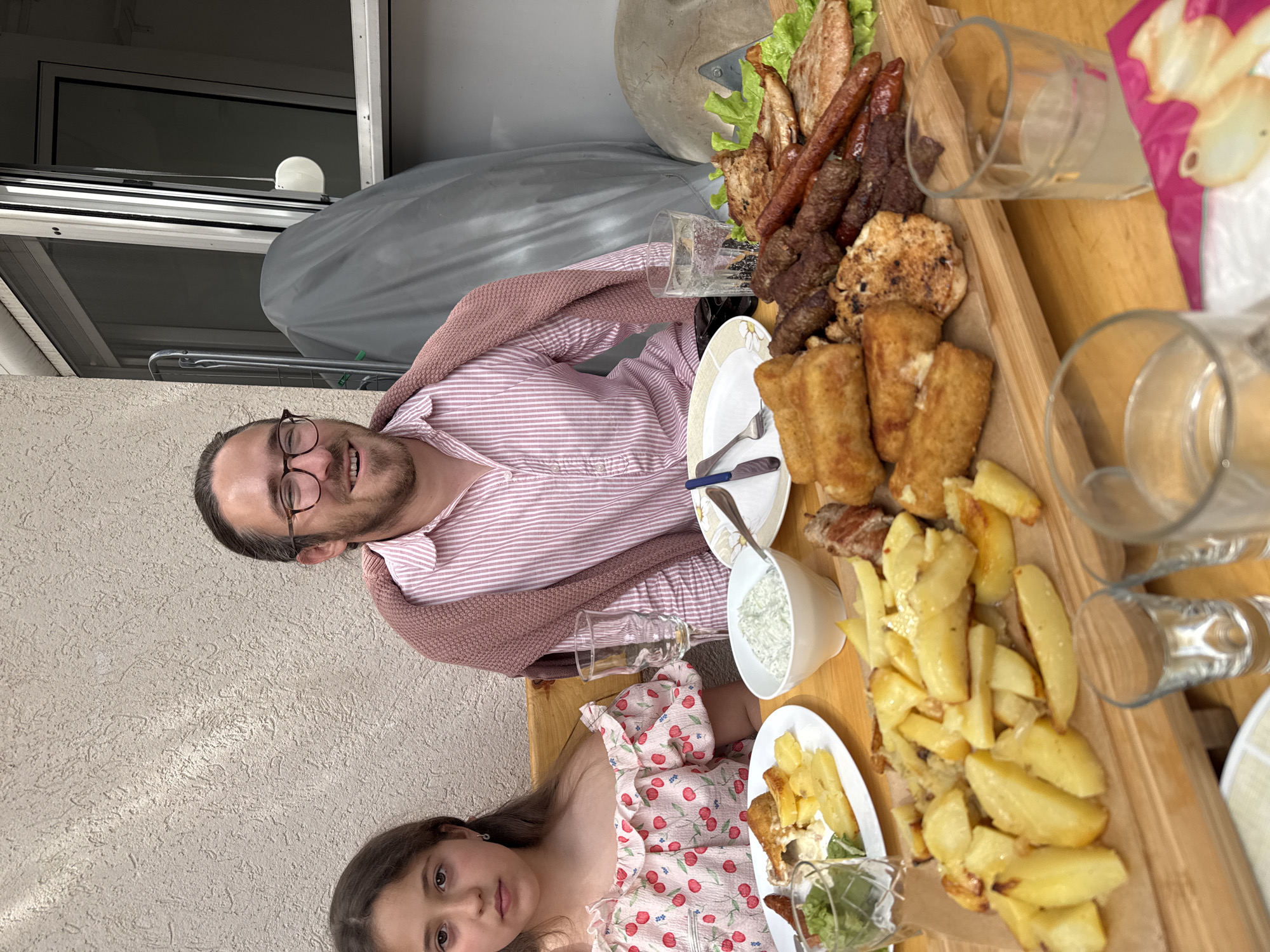 The image depicts a man and a girl seated at a table laden with food. The food includes an array of grilled meats, fried potatoes, and sauces, suggesting a barbecue or outdoor meal. The man is wearing glasses and a pink striped shirt with a sweater draped over his shoulders. The girl is wearing a white dress with a red cherry pattern.
The background features a light-colored exterior wall and a covered grill, suggesting the location is a patio or outdoor dining area. The lack of distinct city features makes it difficult to confirm the specific location in Petrovac na Moru, Montenegro, based solely on the image.