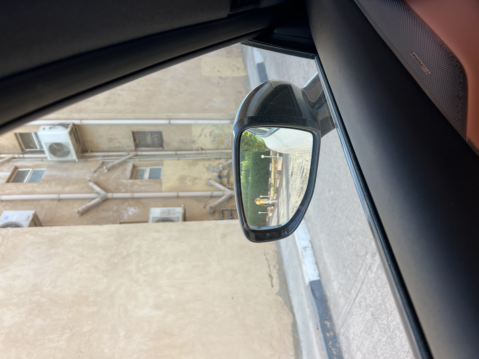 An eye-level, medium shot captures a car's side-view mirror reflecting a person walking on a sidewalk in front of a light-brown building. The building has several windows and air conditioning units visible, along with downspouts and some piping. The sidewalk is bordered by a manicured green hedge. The car interior, with a dark-colored dashboard and a hint of a tan seat, frames the scene. The car is on a paved street with a grey and white curb. The scene is likely in an urban area.