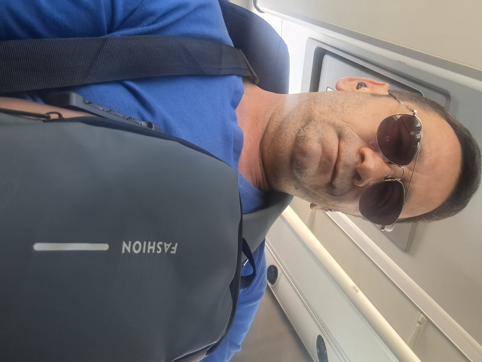 A man is seen from a low angle, wearing sunglasses and a blue t-shirt. He has a grey sling bag with the word "FASHION" printed on it. He is wearing earbuds. The background appears to be the interior of a vehicle, possibly a bus or train, with overhead lighting and white panels. There is no specific information in the image that would indicate the location in Be'er Sheva, Israel.