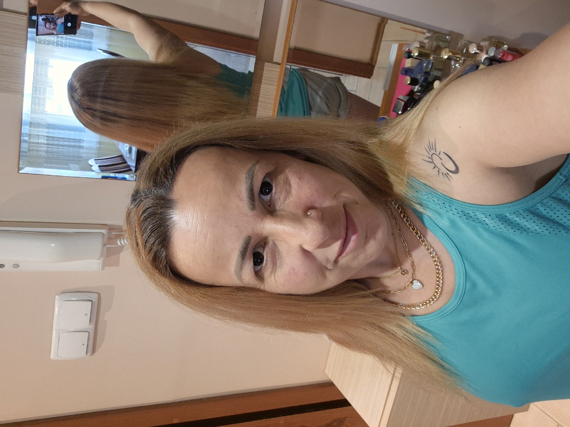 A woman with blonde hair and light skin takes a selfie in a room. She is wearing a teal tank top and multiple necklaces. She has a tattoo of a sun-like symbol on her left shoulder. In the background, a mirror reflects the woman and the room, which includes a dresser with various bottles and a window. A phone is visible in the mirror, held by the woman as she takes the picture. Light switches and an intercom are mounted on the wall to the left of the woman.