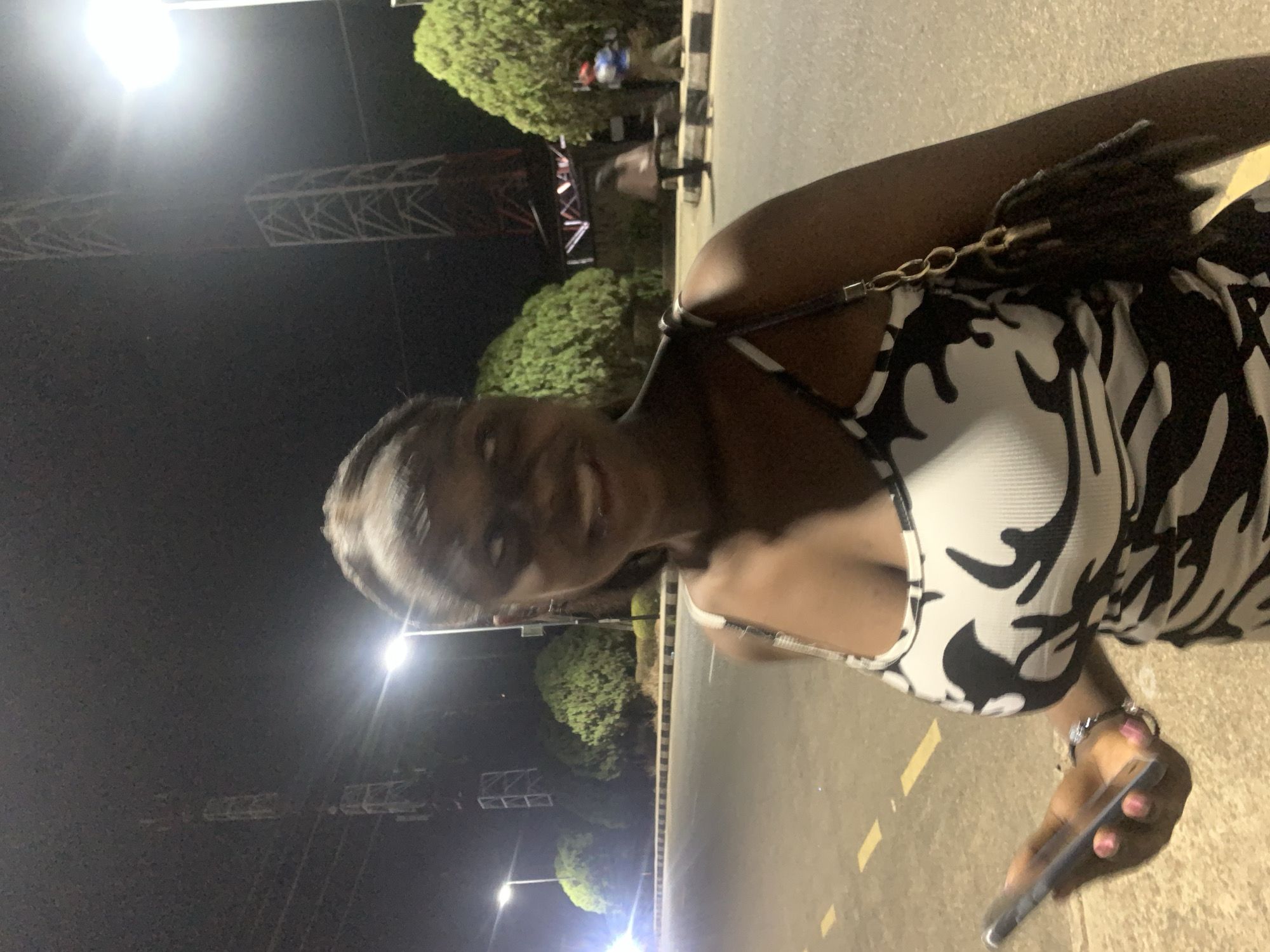 The image shows a person standing on a road at night in Jos, Nigeria. The person is wearing a black and white patterned top and holding a phone. Streetlights illuminate the scene, and trees line the road. A tower-like structure is visible in the background. The person is smiling and looking at the camera.