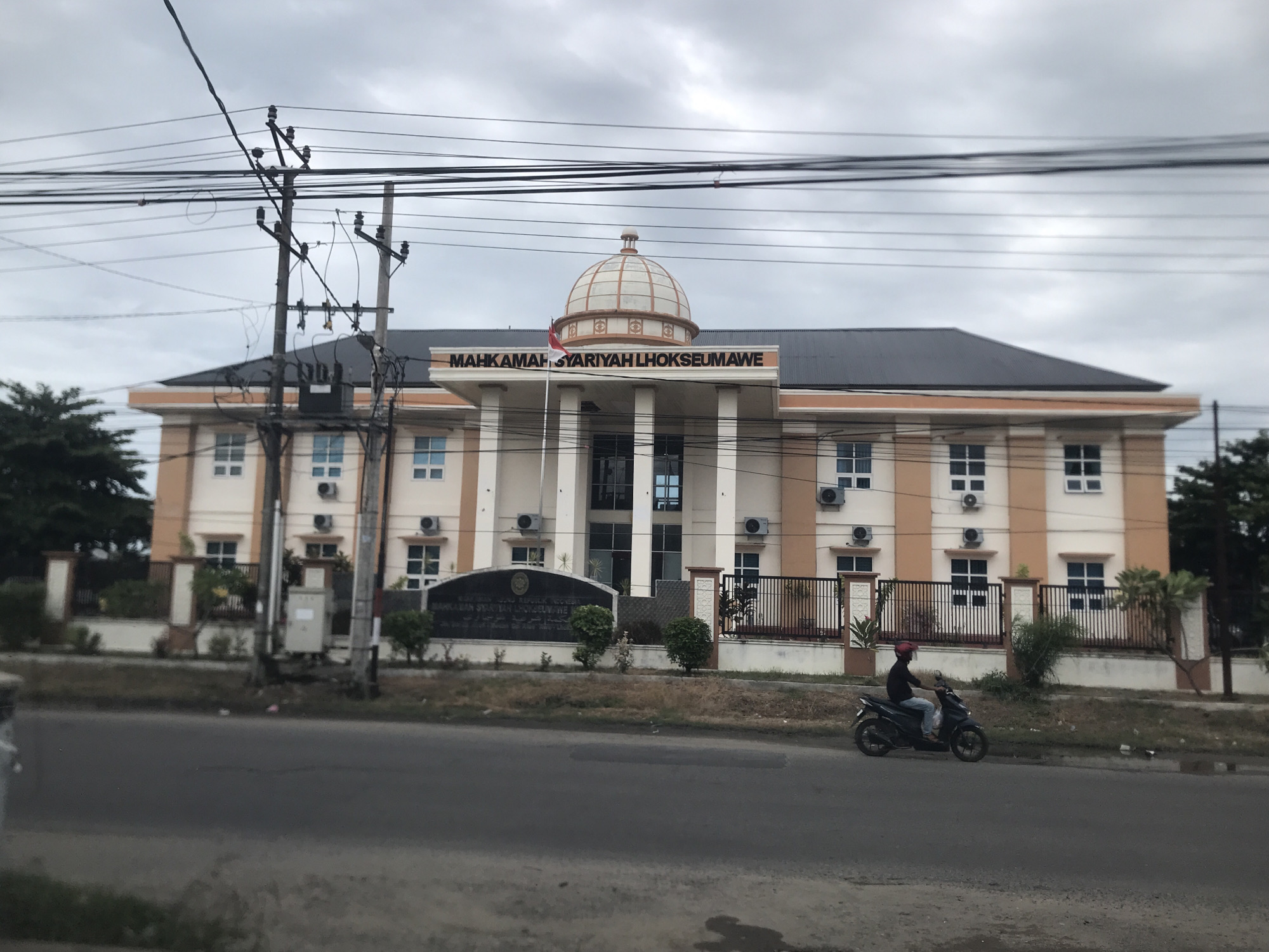 The image shows the Mahkamah Syari'ah Lhokseumawe, a Sharia court in Lhokseumawe, Indonesia. The two-story building is cream-colored with brown trim and a dome in the center. The name of the court is displayed in large letters above the entrance. A motorcycle is driving on the road in front of the building, moving from right to left. There is a pole with power lines to the left of the building. The sky is overcast.
