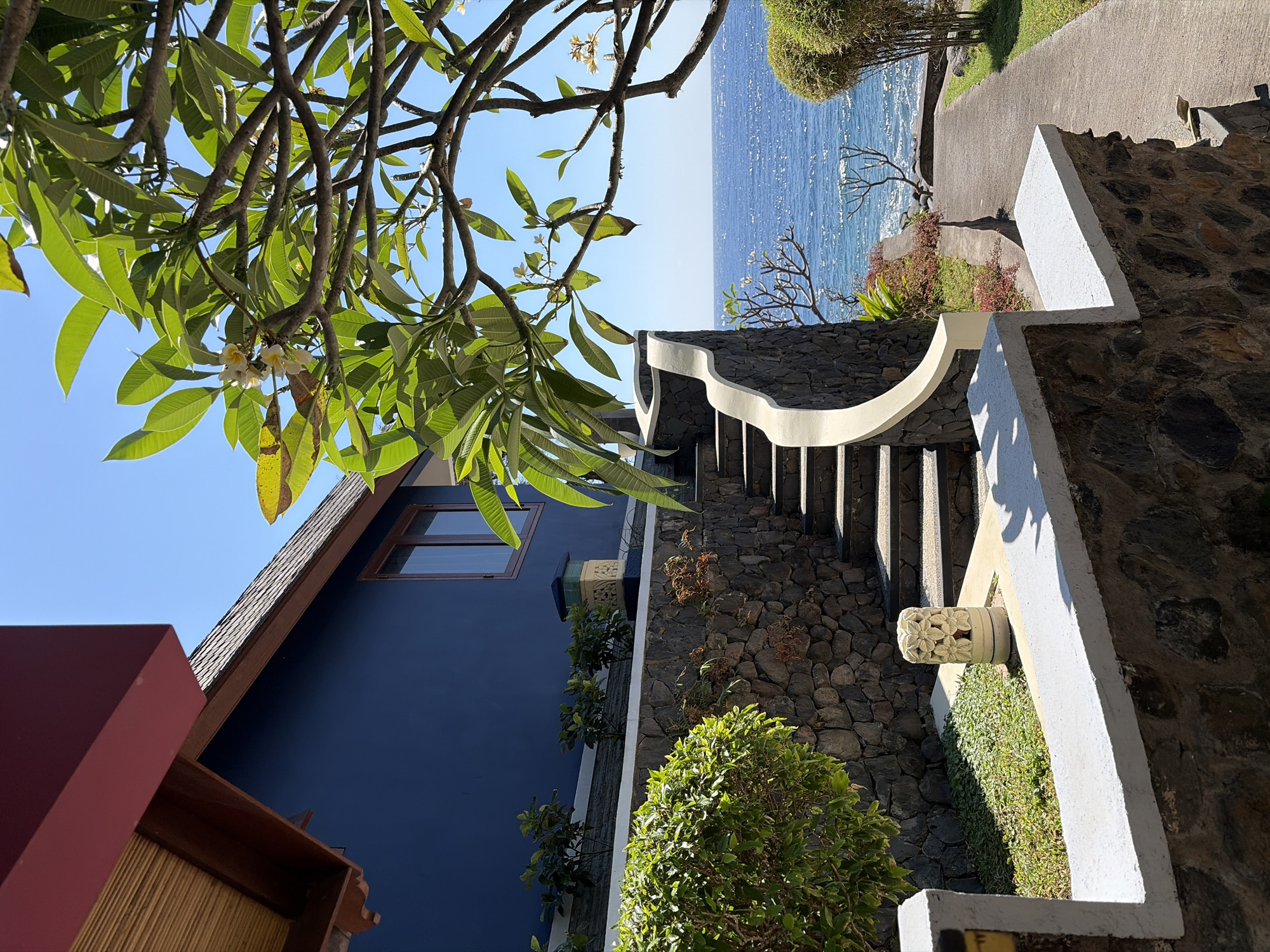 The image depicts an outdoor scene in Abang, Indonesia, likely at a resort or private residence with ocean views. The foreground features a stone wall topped with a white concrete railing, partially obscuring a small garden area with green ground cover and a decorative stone pillar with floral carvings.
To the left, a dark blue building with a wooden-framed window and a dark red overhanging eave is visible. The building sits on a raised platform behind a stone retaining wall. A walkway paved with concrete leads from the foreground towards the ocean on the right.
In the center, a curved stone staircase with a white railing ascends, bordered by a dark stone wall. Lush green foliage from a tree with light-colored flowers overhangs the top of the stairs and extends into the right side of the frame.
The background is dominated by a clear, bright blue sky and the vast expanse of the ocean, its surface sparkling under the sunlight. The shoreline is visible in the distance, with a line of green vegetation and trees along the edge. The overall impression is one of a serene, tropical environment. There are no people visible in the image.
