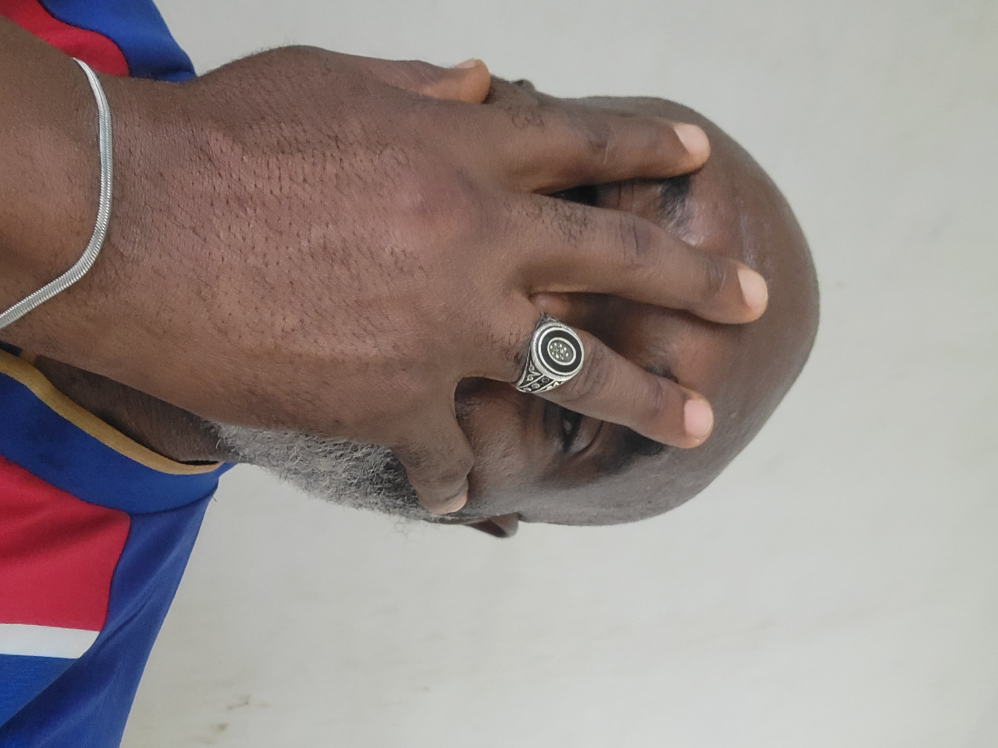 A medium shot shows a dark-skinned man with gray facial hair covering the right side of his face with his right hand. The man has a silver ring on his right ring finger. A silver chain is visible on his upper left arm. The man's body is turned to the left and his face is obscured by his hand. The man is wearing a shirt with blue, red, white, and yellow on the upper left. The background is a plain white wall. This image does not contain enough information to determine a specific location in Maiburiji, Nigeria.