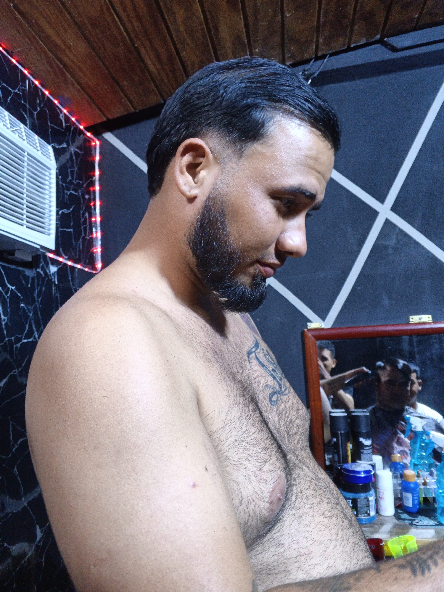 A shirtless man with dark hair and a beard stands indoors, possibly in a barber shop, as suggested by the mirror reflecting a person cutting another's hair. The man has tattoos on his left arm and chest. Behind him is a wall with black marble-like patterns and LED lighting. There's an air conditioning unit on the wall and a wooden ceiling above. There are bottles of hair care products on the table below the mirror. The location is presumed to be in Altagracia, Venezuela, based on external information. The man appears to be looking down.