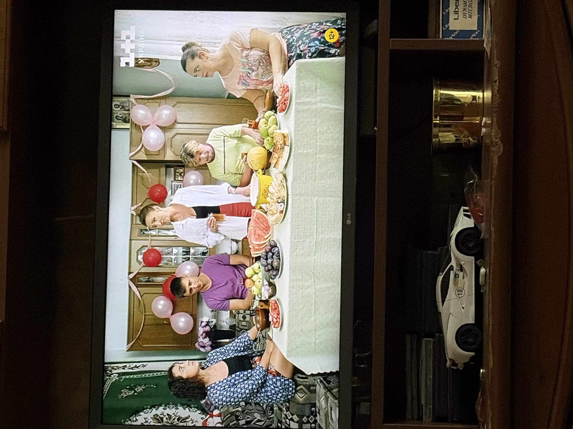 A television screen displays a scene from a television show. In the center of the screen, a group of five people, three women and two men, are gathered around a table laden with food. They appear to be in a room decorated with pink and red balloons. The people are dressed casually, and the food on the table includes fruit, watermelon, and pies. The television itself is mounted above a shelf where several objects are placed, including a white toy car, two gold containers, and a box with text. The background of the room in the show appears to be a domestic setting.