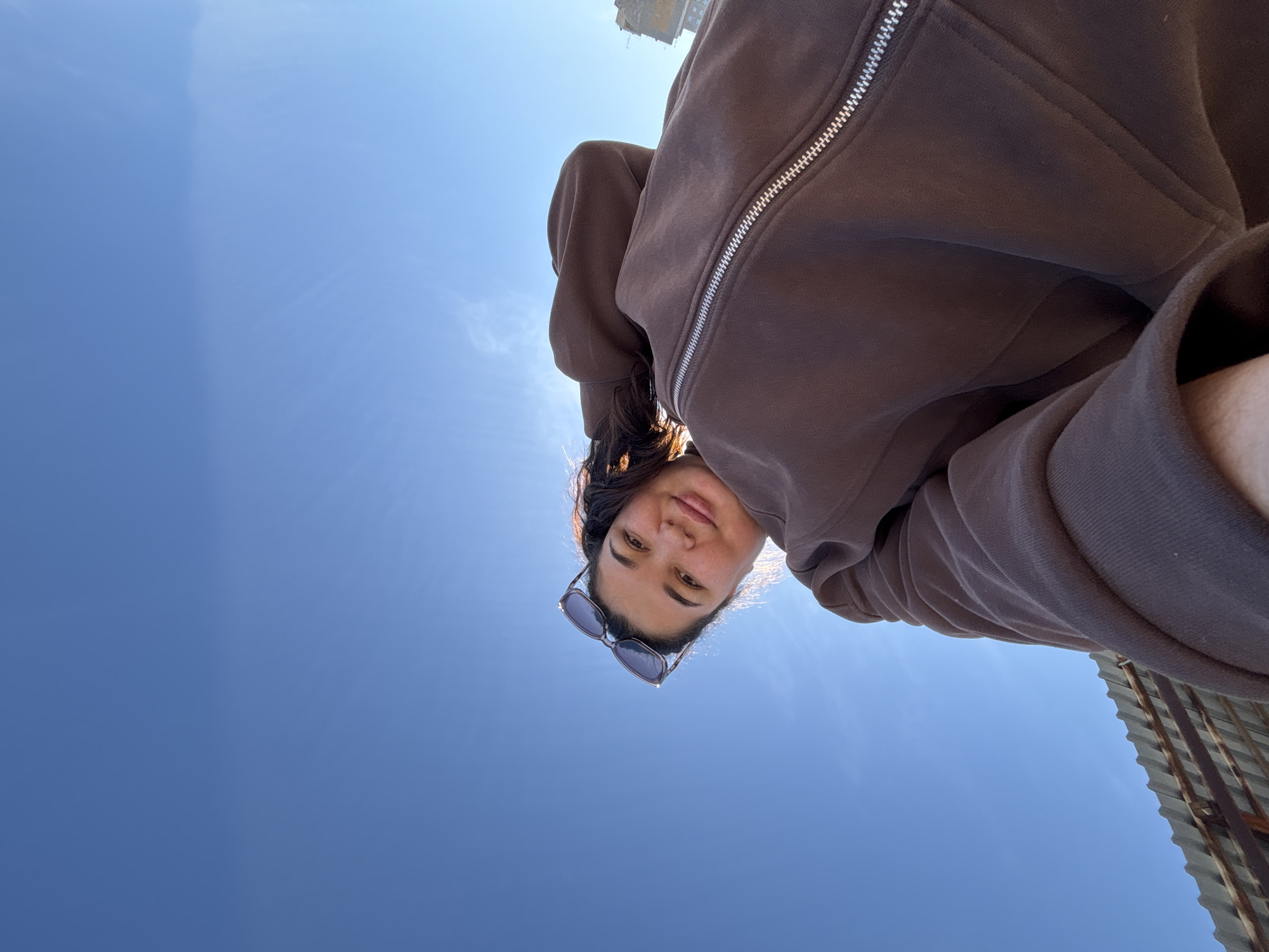 A low-angle selfie photograph shows a woman with dark hair wearing sunglasses and a brown hooded jacket. The background is a clear, bright blue sky. A building is slightly visible in the upper center of the frame. A metal structure is visible in the lower right corner. The location is likely Chaikovsky, Russia.