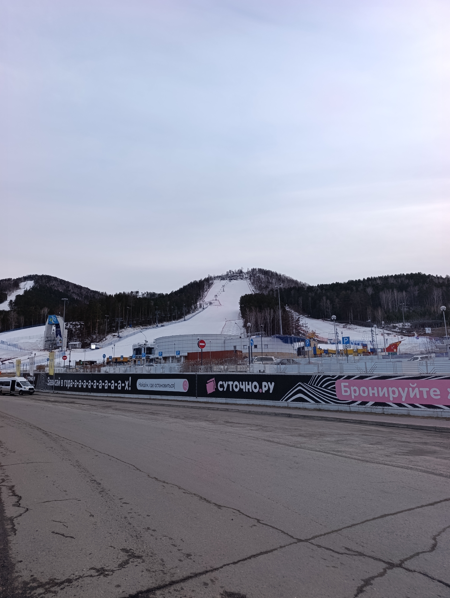 A panoramic view presents the Bobrovy Log Funpark in Krasnoyarsk, Russia. The funpark, identifiable by its ski slopes covered in snow, ascends into a forested hill. In the foreground, a road exhibits signs of wear with visible cracks. Above the road, a long advertising banner is displayed, bearing a message in Russian: "Зависай в гора-а-а-а-а-а-а-а-а-а-х! Найдём, где остановиться," with the website "СУТОЧНО.РУ" and the call to action "Бронируйте" (Book). Several buildings and parking areas are situated at the base of the ski slopes, with vehicles visible, indicating activity at the site. The sky is overcast, suggesting a cold or winter day.