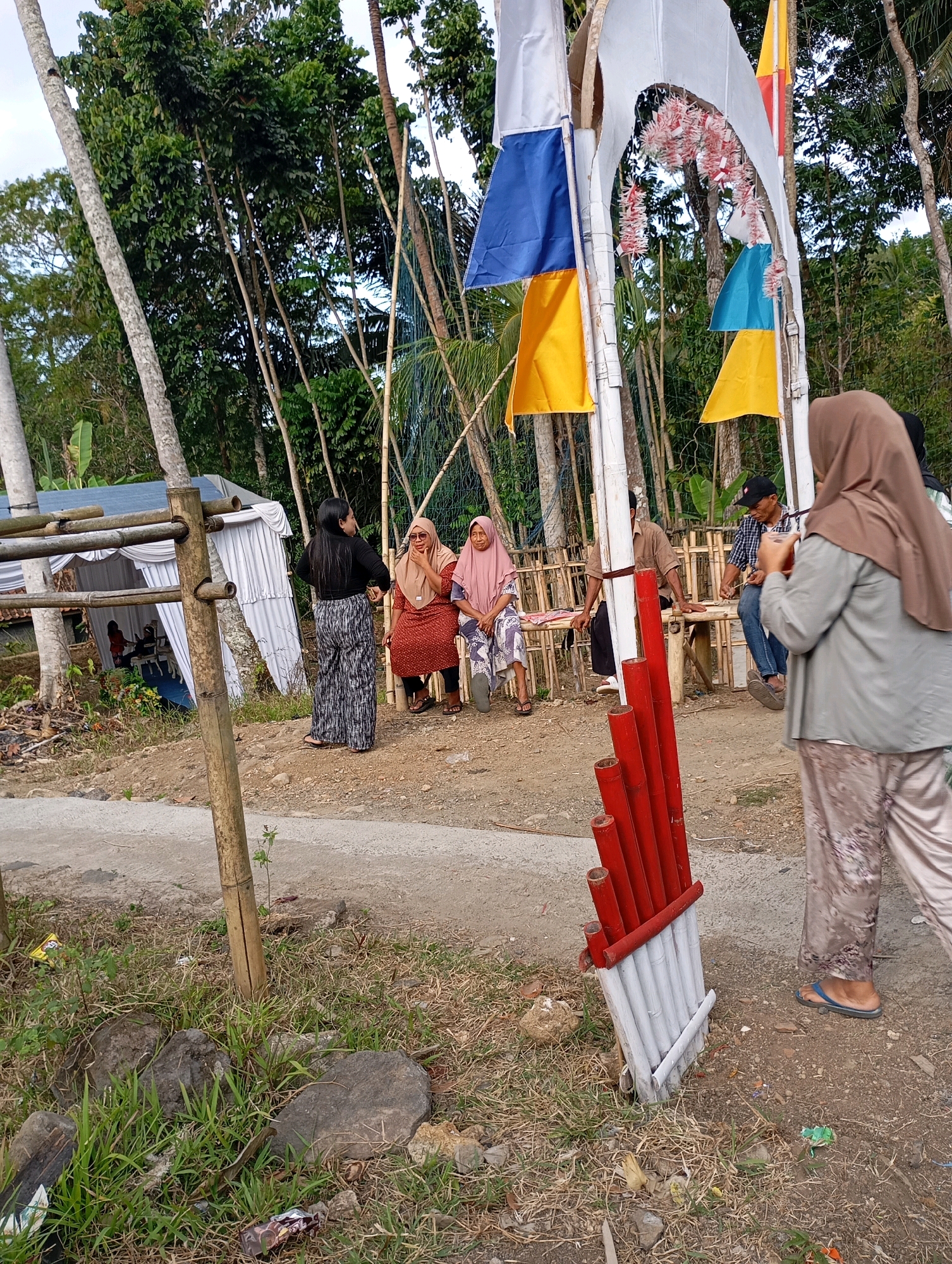 A group of people are gathered outdoors in an area with trees and a dirt path. In the foreground, to the right, a person wearing a tan hijab and patterned pants walks away from the camera. To the left, a bamboo structure is visible, with a section of red pipes attached to it. Behind this, a white tent is set up. Several other individuals are seated and standing in the middle ground. Some are sitting on a wooden bench, and two women are standing near the tent. The area appears to be a rural setting, possibly in Cigunung, Indonesia, given the presence of tropical vegetation. Flags of white, blue, and yellow are displayed on a white archway behind the group. The ground is covered with dirt and some dry grass and rocks.