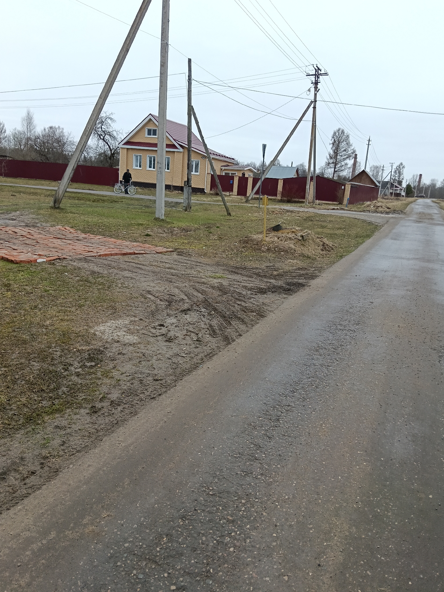 The image depicts a street scene in Красный Маяк, Russia, on an overcast day. The road is paved and wet, suggesting recent rain. A residential area lines the street, featuring houses with brick walls and fences. Utility poles are visible along the roadside, supporting overhead wires. A person is riding a bicycle on the sidewalk in front of one of the houses. Construction materials, specifically bricks, are piled on the ground near the roadside. The environment consists of grass and bare patches of earth, typical of a rural or suburban setting.