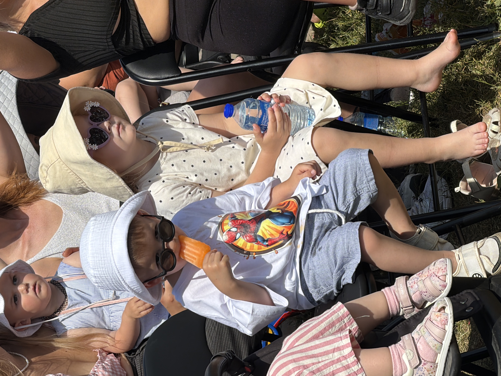 The image depicts an outdoor daytime scene in Khimki, Russia, featuring several individuals, primarily young children and partially visible adults, seated in black foldable chairs on a grassy area under direct sunlight.
In the foreground, two young children are prominent. On the left, a boy wears a white bucket hat, round dark sunglasses, and a white short-sleeved t-shirt featuring a large circular graphic of Spider-Man against an orange and yellow radial background. He is holding an orange popsicle with his right hand and appears to be eating it. His lower body is clad in light blue pinstriped shorts with a visible white drawstring. His feet are mostly obscured, but light-colored footwear is partially visible.
To the boy's right, a girl wears a wide-brimmed light beige sun hat with a chin strap and purple heart-shaped sunglasses adorned with small white daisy decals. She has a light-colored dress or top with dark polka dots. She holds a transparent plastic water bottle with a blue cap in both hands; Cyrillic text, including "СТАРО-МЫТИЩИНСКИЙ", is visible on the bottle's label, consistent with the Moscow Oblast region near Khimki. Her legs are bare, and she is barefoot, with her feet resting near the base of her chair.
Behind the boy, a smaller child with a white bucket hat, a light blue and white striped sleeveless top, and a dark choker necklace is partially visible, looking slightly to the right. Glimpses of several adults are also present, including a woman with reddish-brown hair in a light-colored top behind the smaller child, and a woman in a black ribbed crop top to the right of the girl. Another person's legs in pink and white striped trousers and pink floral sandals are seen in the lower left.
All individuals are seated on black metal foldable chairs. The ground beneath the chairs is covered in dry-looking green grass. Bright ambient light indicates sunny weather conditions.