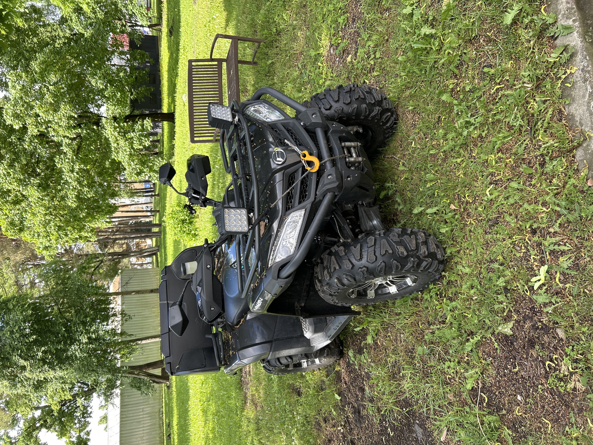 A black all-terrain vehicle (ATV) is parked on a grassy area in a park. The ATV is facing forward and is slightly angled to the right. It has large, knobby tires, a storage box on the back, and multiple LED lights on the front. A wooden park bench is visible to the right of the ATV. In the background, there are several trees and a long, light-colored metal building with a grey roof. The ground around the ATV is covered in green grass and some brown dirt. The scene appears to be a bright, sunny day.