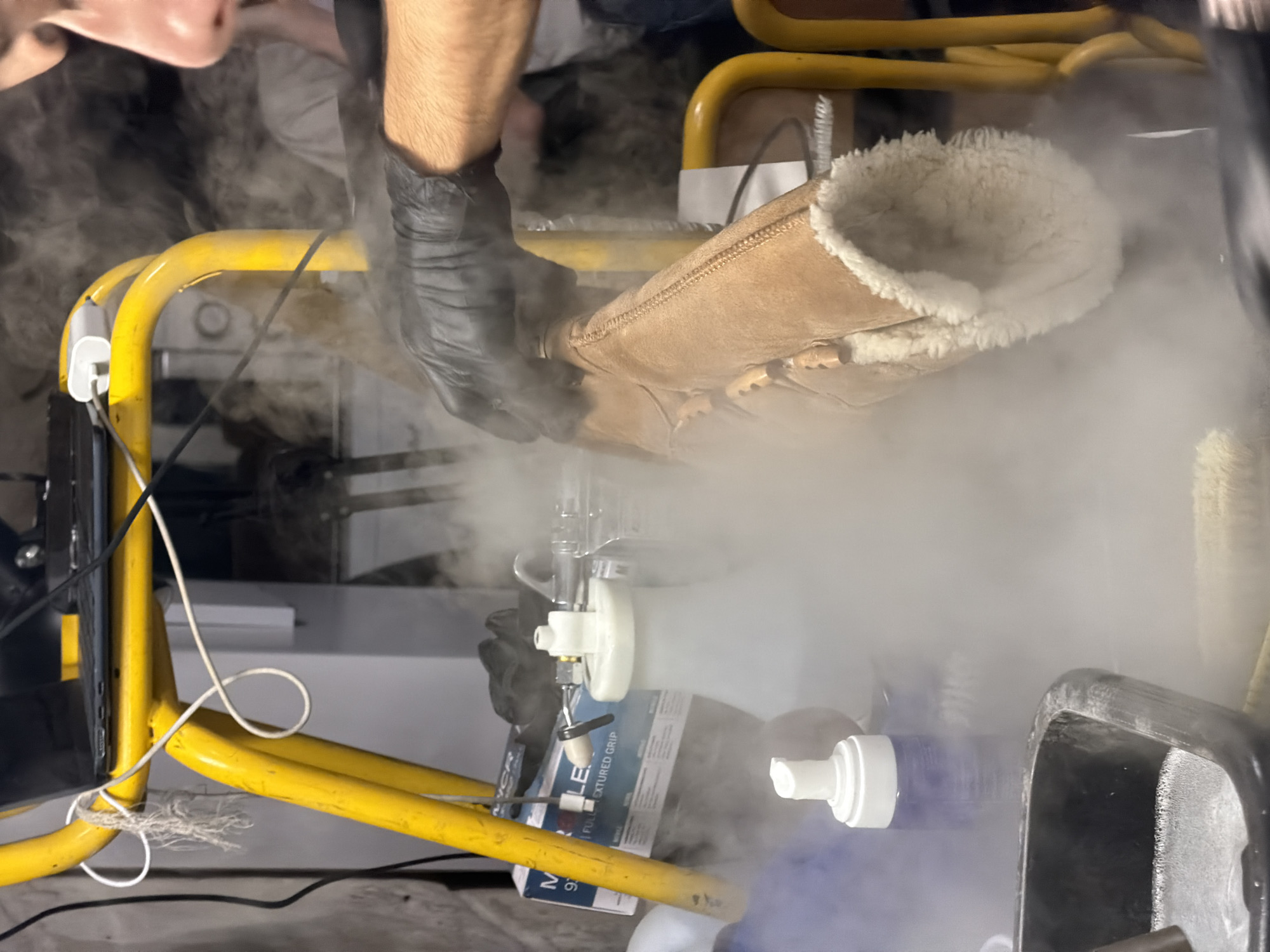 A person wearing black gloves is holding a tan Ugg boot over a device that is emitting a thick white vapor. The device appears to be connected to a system of yellow metal tubing. A box with blue and white packaging is partially visible beneath the device. The environment is cluttered, with a black tray-like object visible in the lower right corner. The location is unknown, and there is no indication it is in Paris, France.