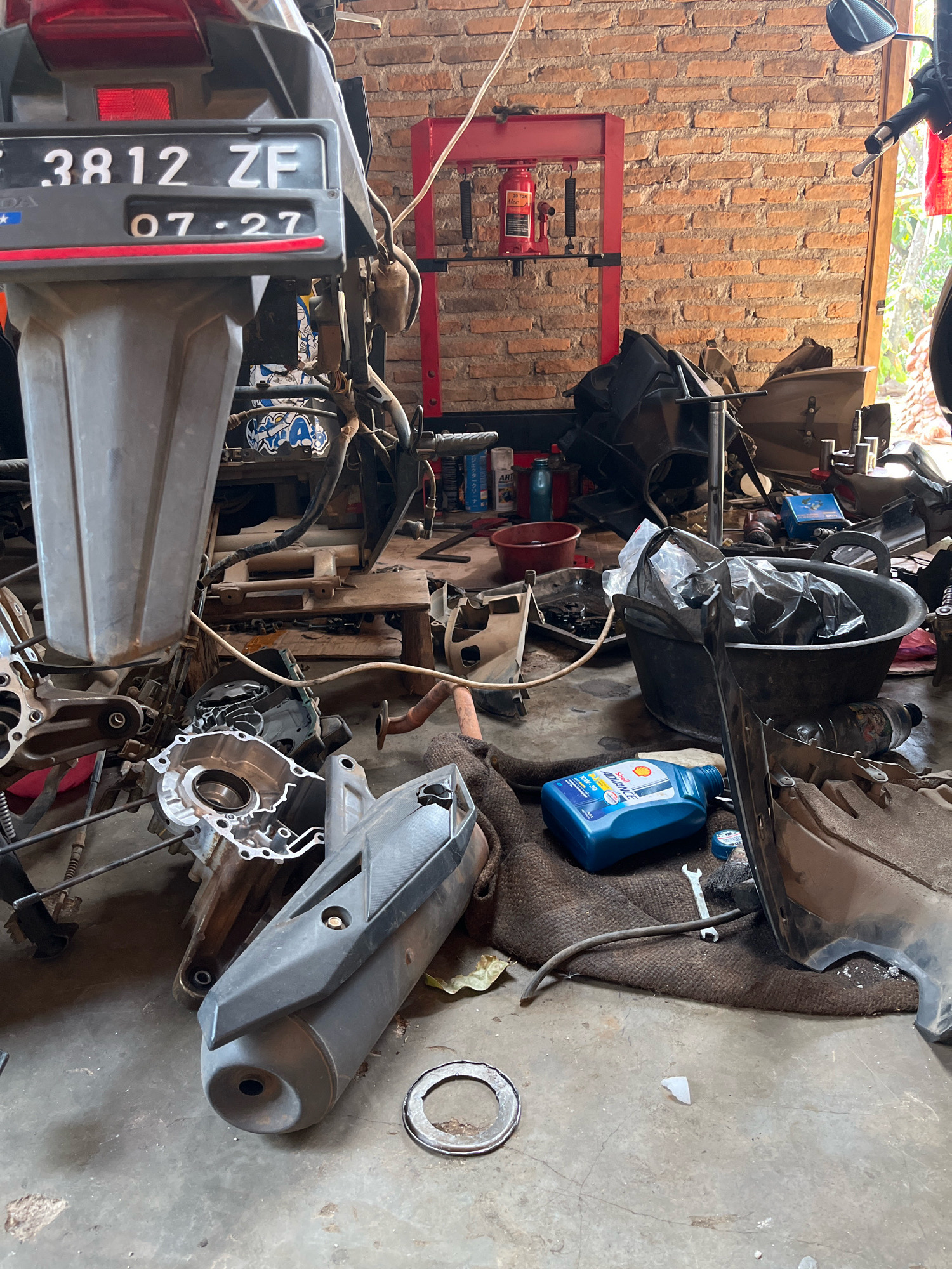 The image depicts a cluttered motorcycle repair area within a building with a brick wall. A motorcycle's rear section with a license plate reading "3812 ZF 07-27" is visible at the top left. Various motorcycle parts, including an exhaust pipe, engine components, and plastic fairings, are strewn across the concrete floor and on a brown mat. A large black tub filled with black plastic bags and a blue bottle of Shell Advance motorcycle oil are also present. In the background, a red hydraulic press stands against the brick wall, and some shelves with bottles and other items are visible. The scene appears to be an indoor workshop or garage.