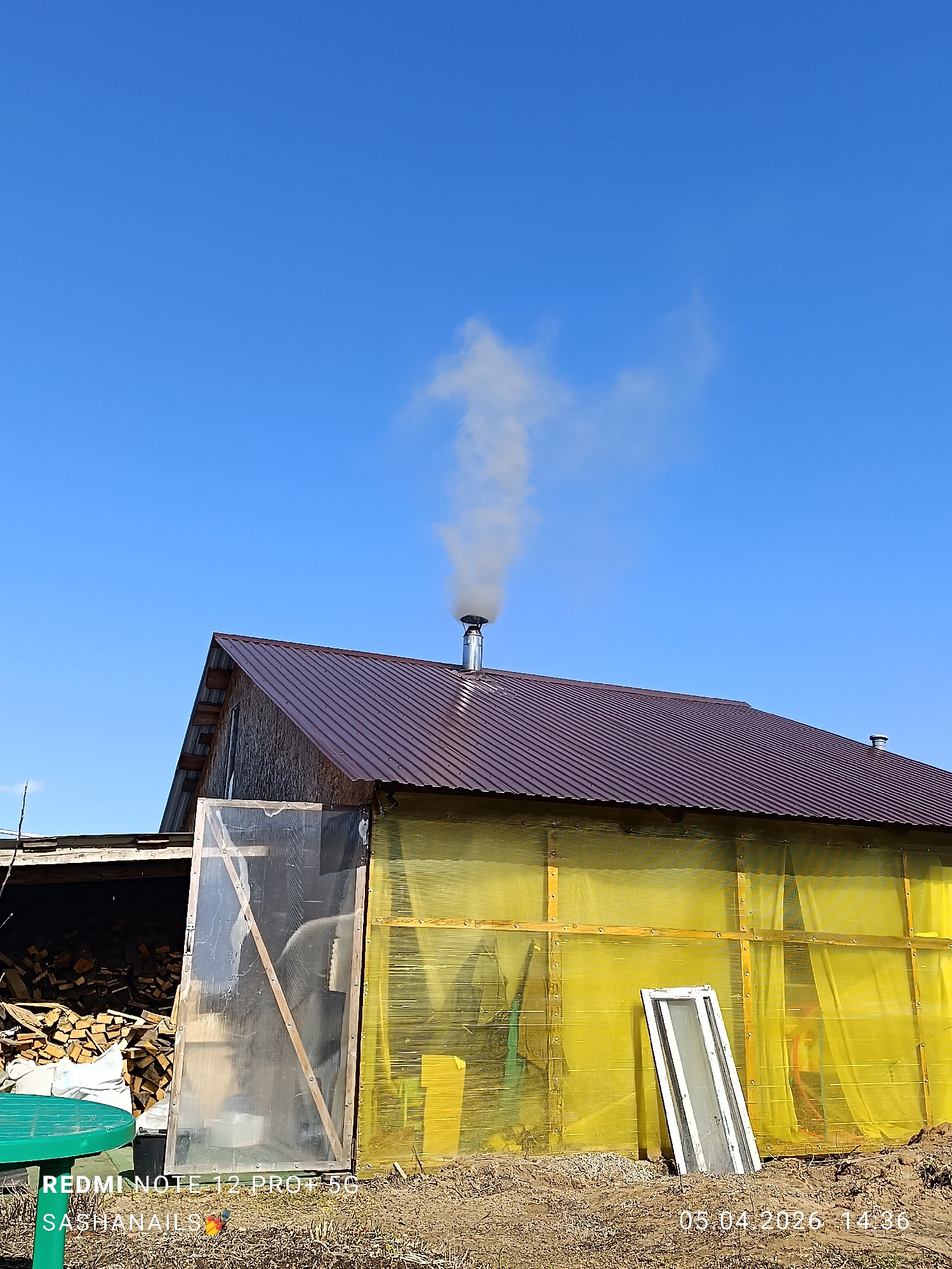 A photograph captures a rural scene, dominated by a building with a dark brown corrugated metal roof. A silver chimney rises from the roof, emitting a plume of grey smoke into a clear, cloudless blue sky. The building's walls are covered in translucent yellow plastic sheeting, revealing a wooden frame underneath. Leaning against one wall is a white-framed window. To the left of the building is a partially visible wood shed with a pile of firewood. In the foreground, a green plastic table is visible, along with some construction materials. The scene is located in Chaikovsky, Russia. The photograph includes a watermark from a Redmi Note 12 Pro+ 5G mobile phone, indicating the device used to capture the image. The watermark also displays the date "05.04.2026 14:36," indicating the date and time the photo was taken.