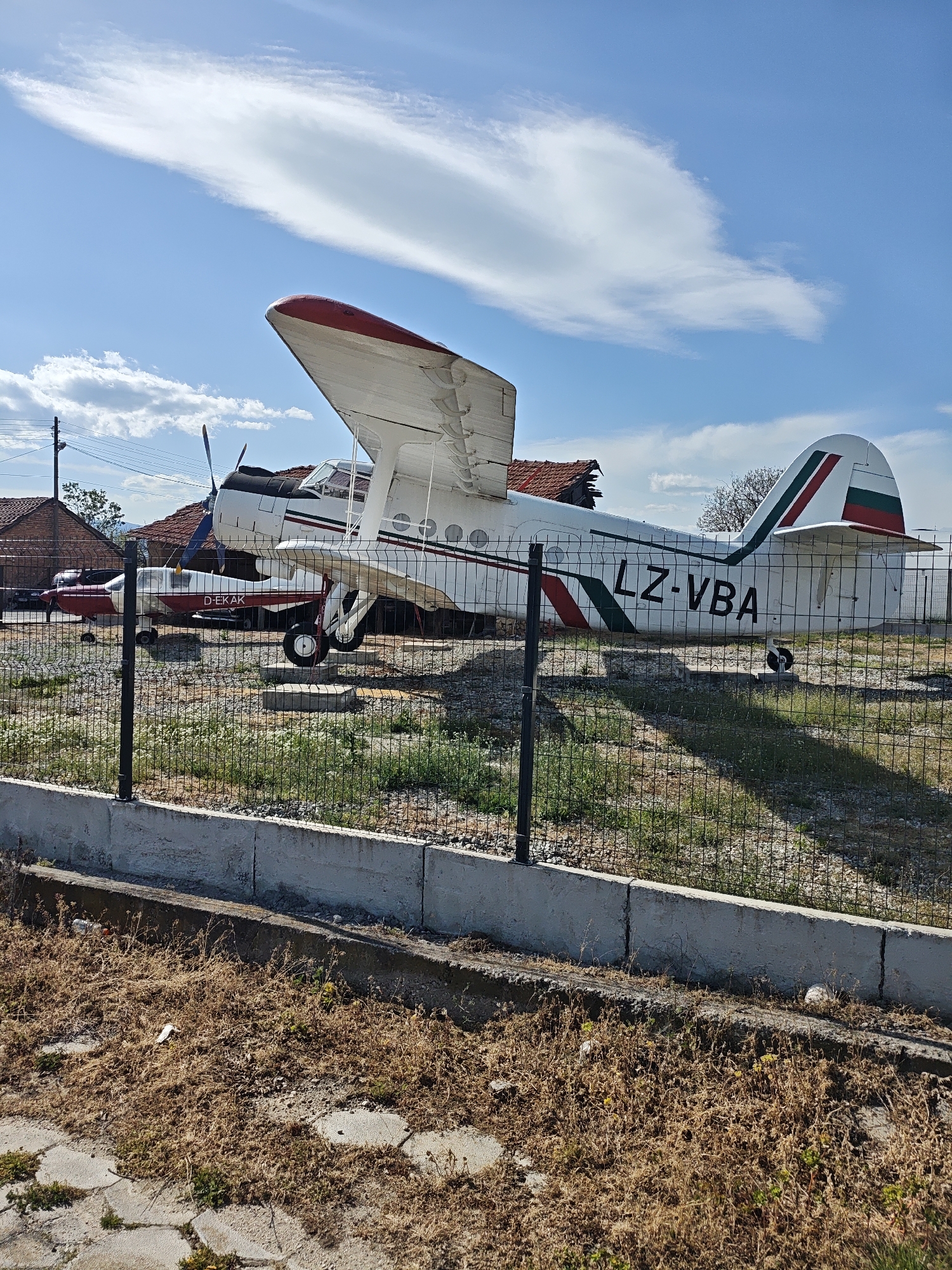 The image shows an Antonov An-2 airplane, registered as LZ-VBA, behind a chain-link fence and a low concrete wall. The biplane is painted white with red and green stripes, and the tail markings suggest it's from Bulgaria. Another smaller plane, registered as D-EKAK, is parked nearby. Both airplanes are in an outdoor area with grass and weeds growing. In the background, there are houses with red tiled roofs. The sky is blue with scattered white clouds. The location is in the city of Unknown, Unknown.