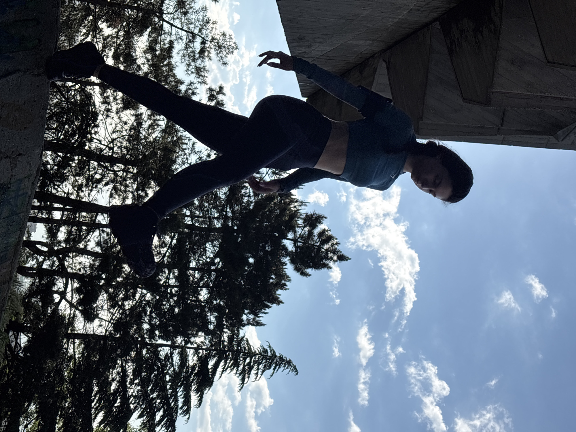 This media file displays a dynamically posed individual captured from an unusual, rotated perspective, where the subject appears to be defying gravity. A person, dressed in dark athletic wear including a long-sleeved crop top and leggings, is positioned horizontally across the frame. Their left leg is extended straight, with the foot pressed against a dark, textured vertical surface that could be a tree trunk or concrete pillar, while their right leg is bent. The person's torso is slightly arched, and their arms are extended outwards with open hands, suggesting a balancing act or an artistic pose like dance or yoga. Their head is tilted downwards, partially silhouetted.
The background features a bright blue sky filled with scattered white cumulus clouds. On the left side, dense, dark green tree foliage creates a natural silhouette against the sky. On the right, a large, angular concrete structure, possibly part of an underpass or building, casts a dark shadow and partially frames the scene. The strong backlighting emphasizes the silhouette of the subject, the trees, and the structure. The scene is brightly lit, indicating daytime. No visible text is present. This outdoor setting is located in Varna, Bulgaria.