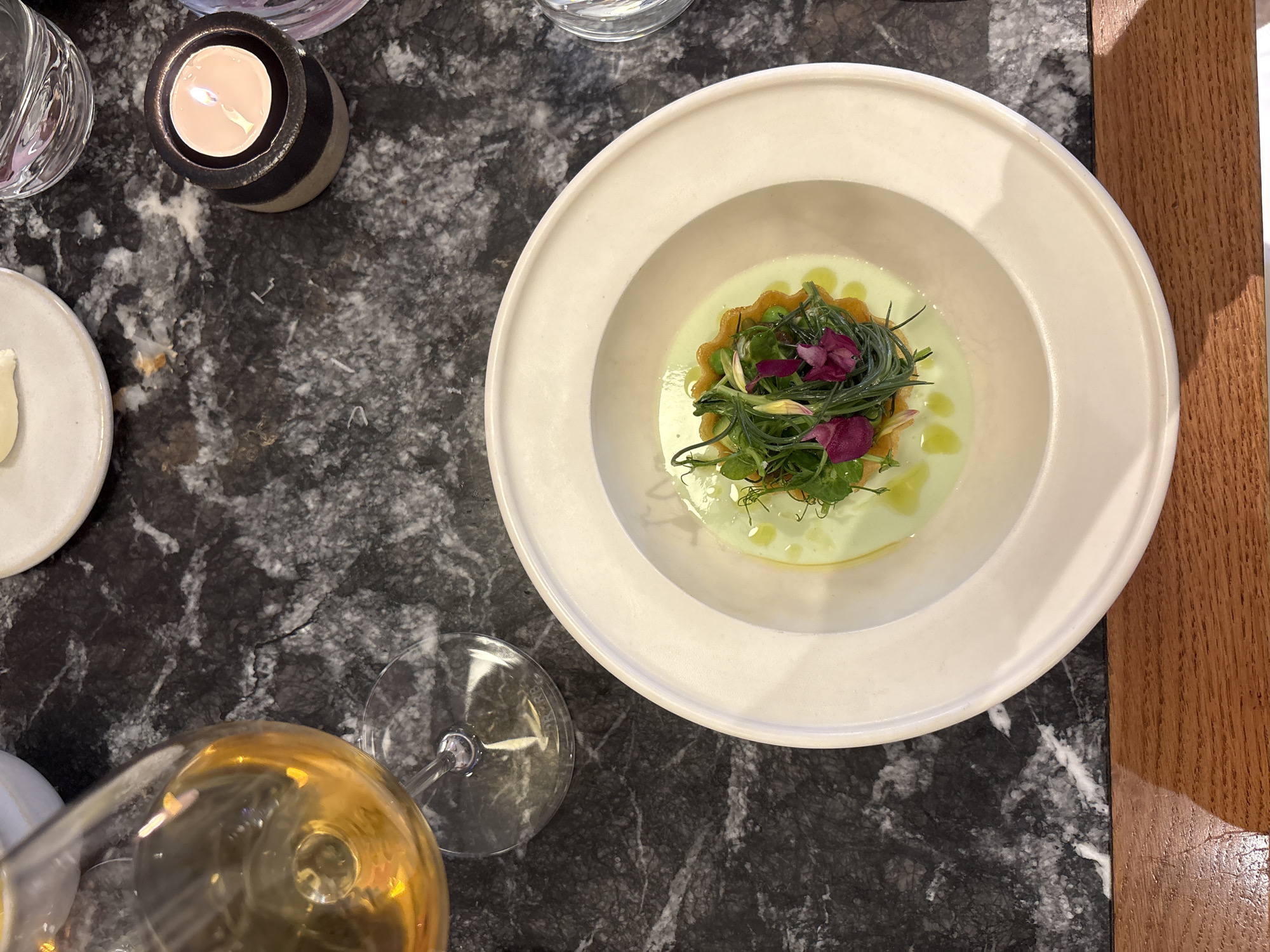 A close-up, overhead shot shows a white, shallow bowl containing a vibrant green soup with a small, intricate garnish of greens and purple flowers. The bowl is placed on a dark, speckled marble tabletop, with a wooden surface visible at the bottom of the frame. To the left, a wine glass with amber-colored liquid is partially visible. To the right, a small, lit candle in a dark holder and a partially obscured water glass are also on the table. The scene appears to be an upscale dining setting. There is no indication of the specific location in London, United Kingdom.