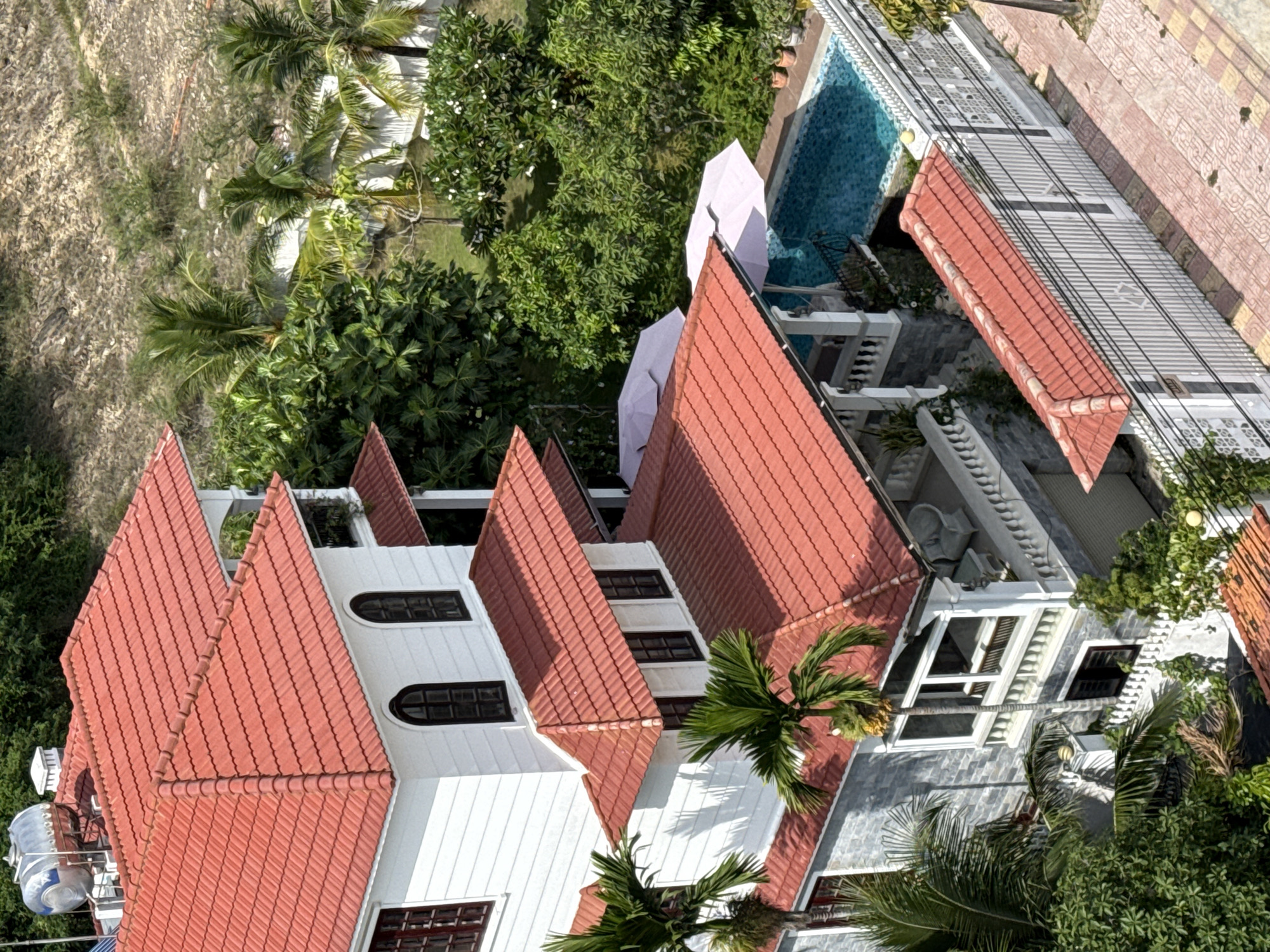 The image captures an aerial view of a multi-story villa with red tile roofing, situated in Bac Nha Trang Ward, Vietnam. The villa features white exterior walls and multiple balconies with white railings. Surrounding the villa are lush green trees, including palm trees, and a swimming pool with clear blue water. Two light-colored umbrellas are positioned near the pool. A white fence encloses the property, and a tiled walkway is visible in the foreground. The overall scene suggests a tropical, residential setting. There are no people visible in the image.