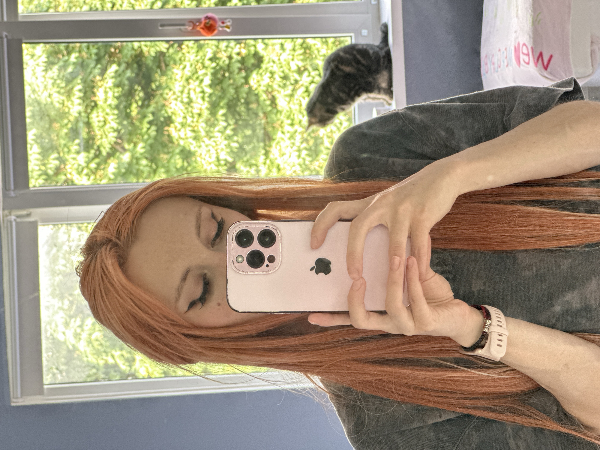 A woman with long, orange hair, wearing a dark grey t-shirt and a light pink smartwatch on her left wrist, takes a selfie in a mirror. She is holding a light pink smartphone in her hands. Her eyes are closed, and she is wearing winged eyeliner. In the background, a window looks out onto lush green trees. On the windowsill, a tabby cat sits facing away from the camera, looking out the window. The room appears to be a bedroom, with a pink and white patterned blanket visible on the right side of the frame.
