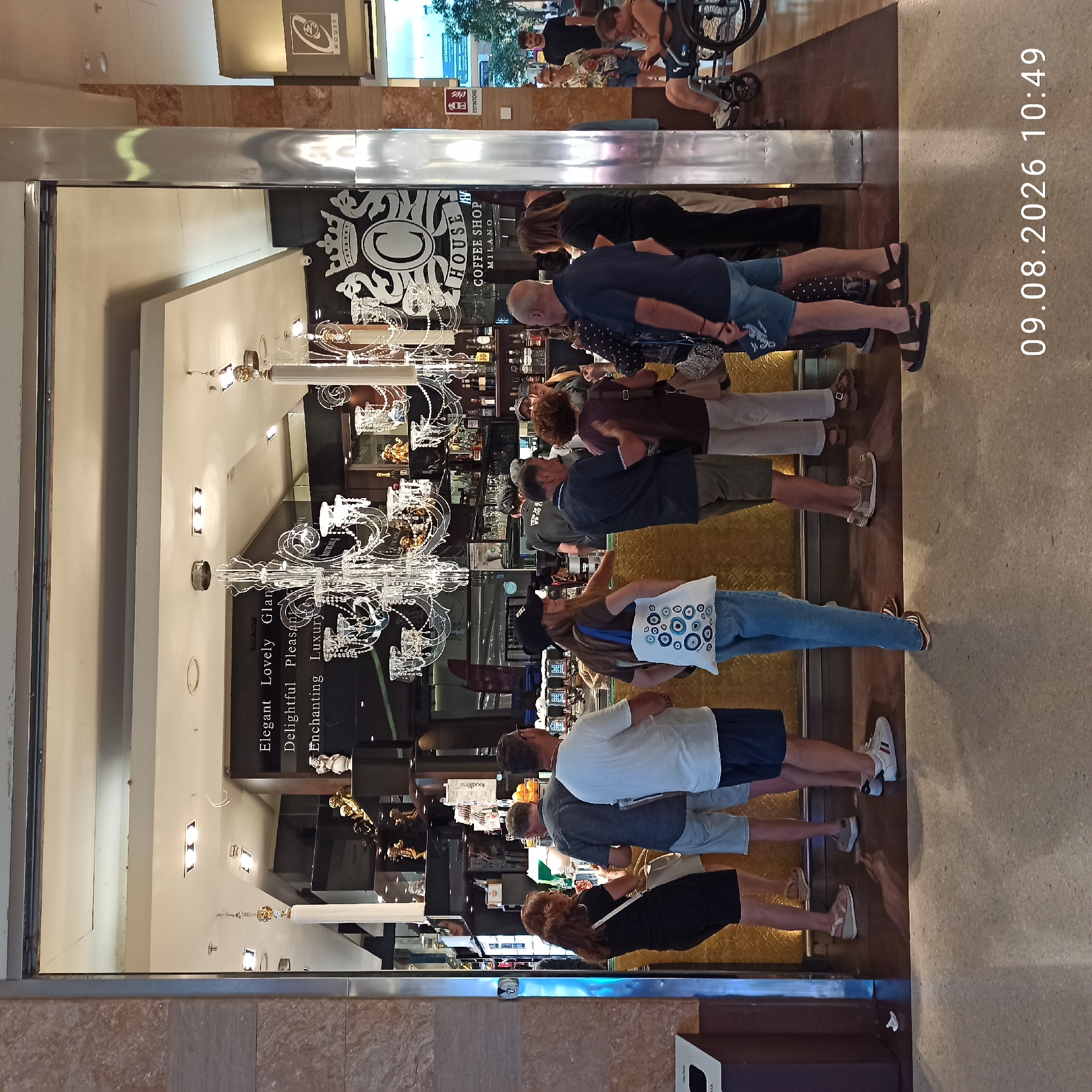 The image shows a street scene in Molfetta, Italy. A group of people are standing in front of a shop with a large chandelier and displays of merchandise. Some people are looking at the merchandise, while others are talking to each other. A woman in a black dress is standing on the left side of the image, looking at the shop. A man in a grey shirt and shorts is standing in the middle of the image, looking at the shop. A man in a white t-shirt and shorts is standing on the right side of the image, looking at the shop. There are also several other people in the background, including a woman in a wheelchair. The shop appears to be a cafe or coffee shop, with a bar and seating area. The shop is decorated with elaborate chandeliers and other decorative items. The street outside the shop is paved with cobblestones. The image is dated 09.08.2026 at 10:49.