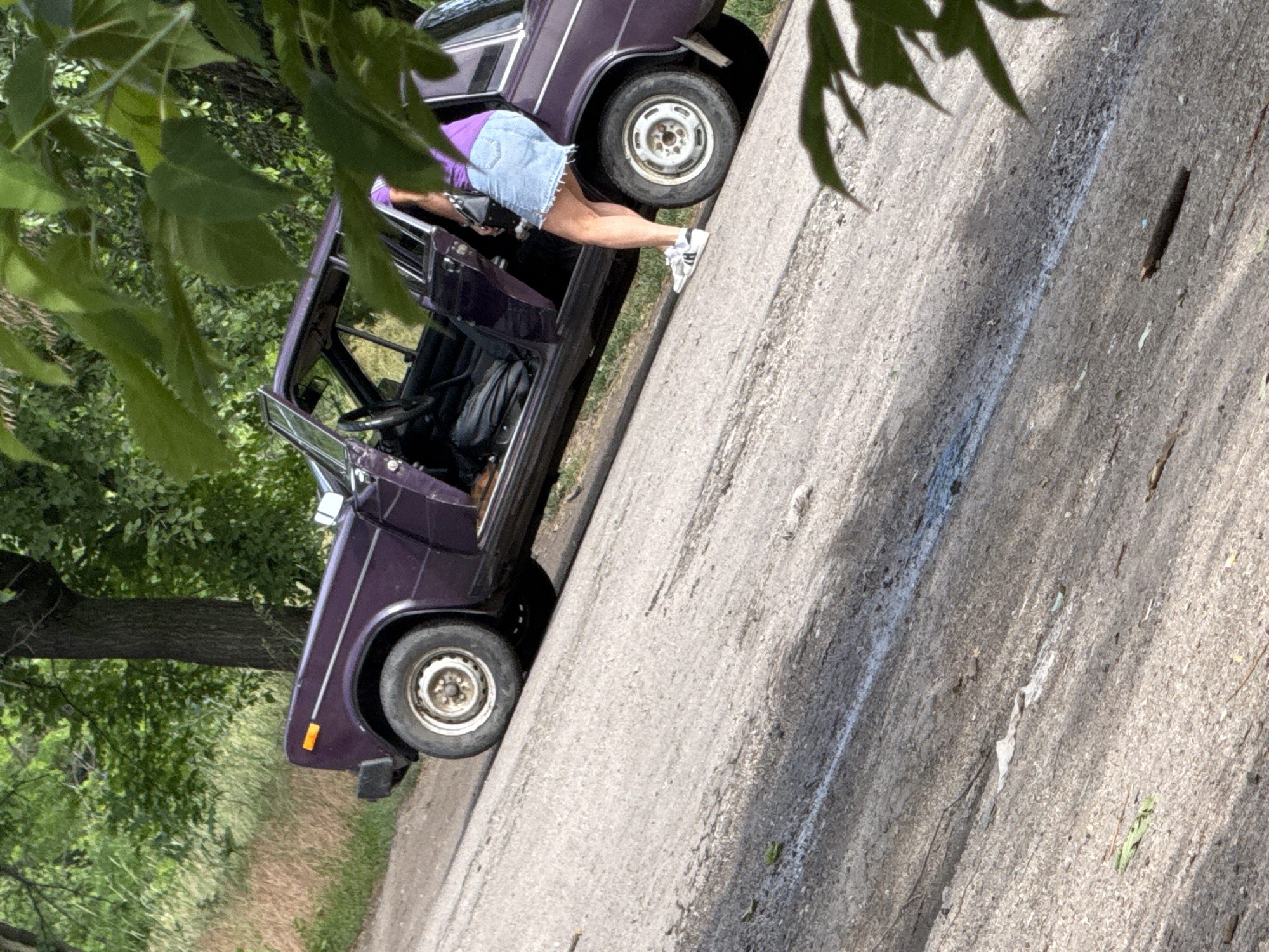 A purple car is parked on the side of a rural road with its driver's side door open. A person wearing a purple shirt and a denim skirt stands next to the open door, appearing to be either entering or exiting the vehicle. The car is old and appears to be in fair condition. The surrounding environment is green and wooded, with trees and foliage visible. The road is paved and shows signs of wear.