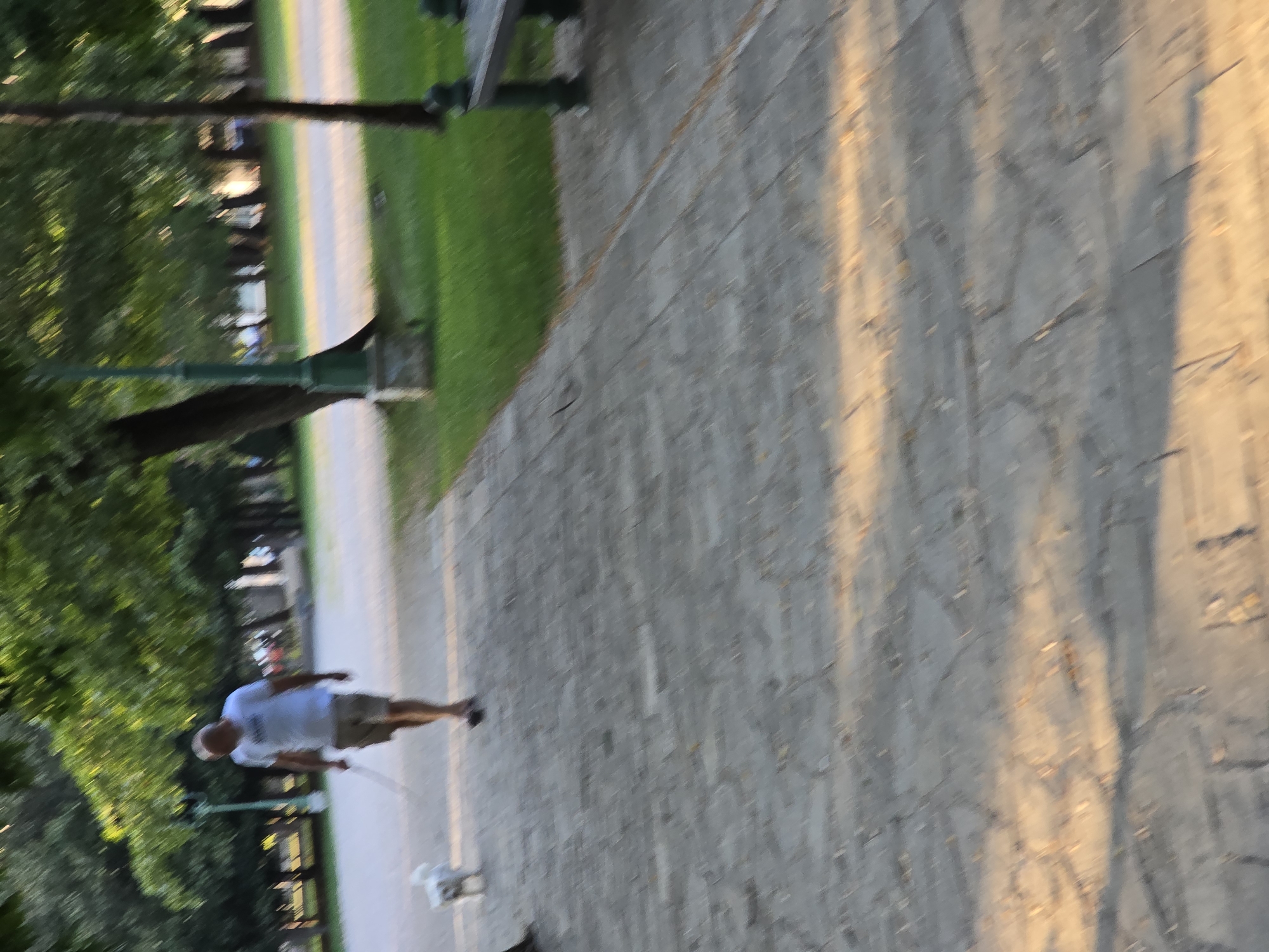 A man in a white t-shirt and khaki shorts is walking a small white dog on a leash along a paved pathway in a park. The pathway is made of light-colored stones. To the right of the pathway is a well-maintained green lawn and a dark bench. Behind the lawn, there are trees with full green foliage, casting shadows on the path. In the background, there is another pathway or road and more trees. A lamppost with a dark green base is visible on the left side of the image. The sun appears to be setting or rising, casting warm light and long shadows across the scene. The image is slightly blurred due to motion. This location is likely within Kruševac Park in Podgorica, Montenegro, given the typical park features and tree types.